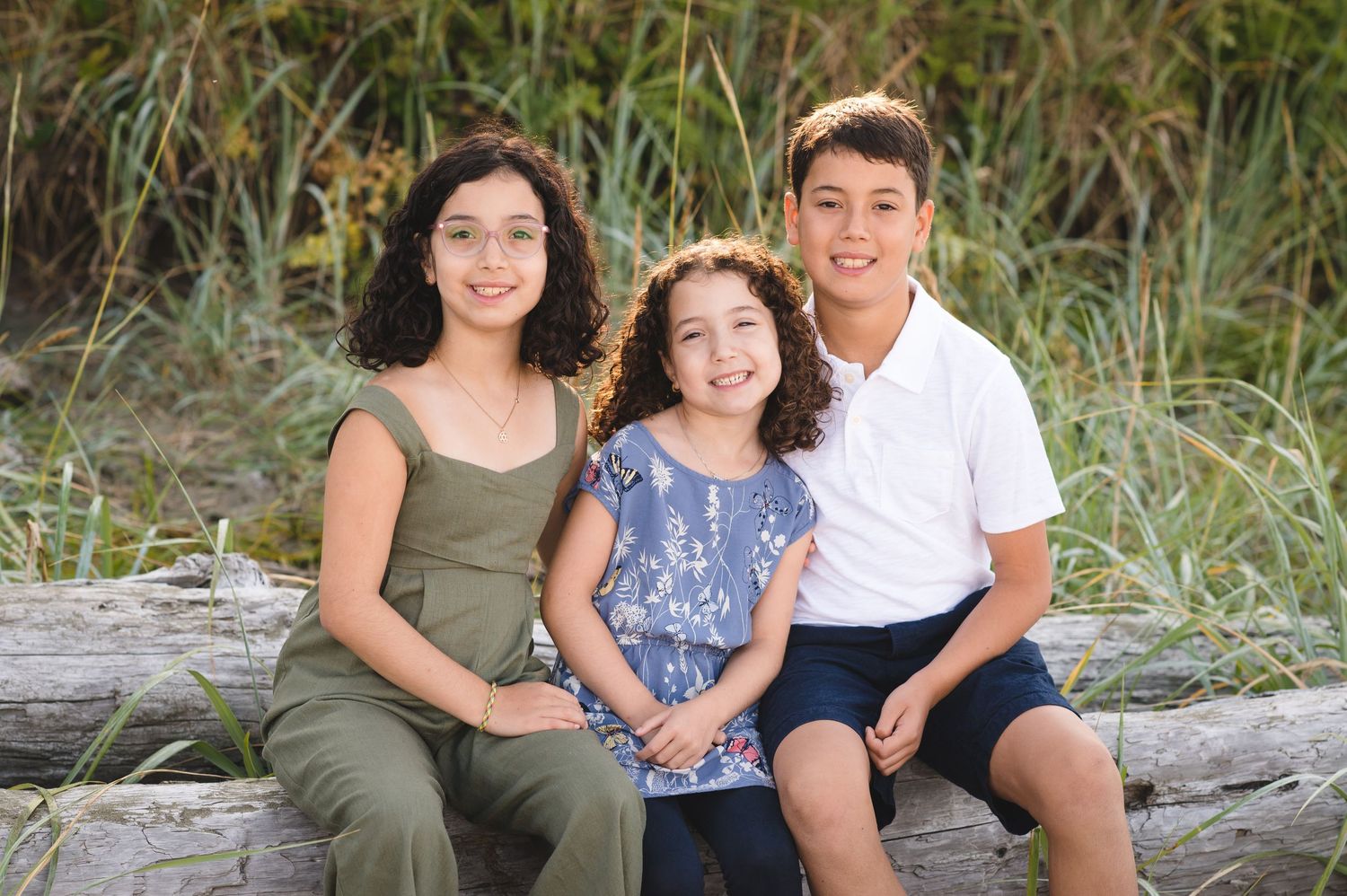 Candid family moment captured on beach logs with sea grass in the background.