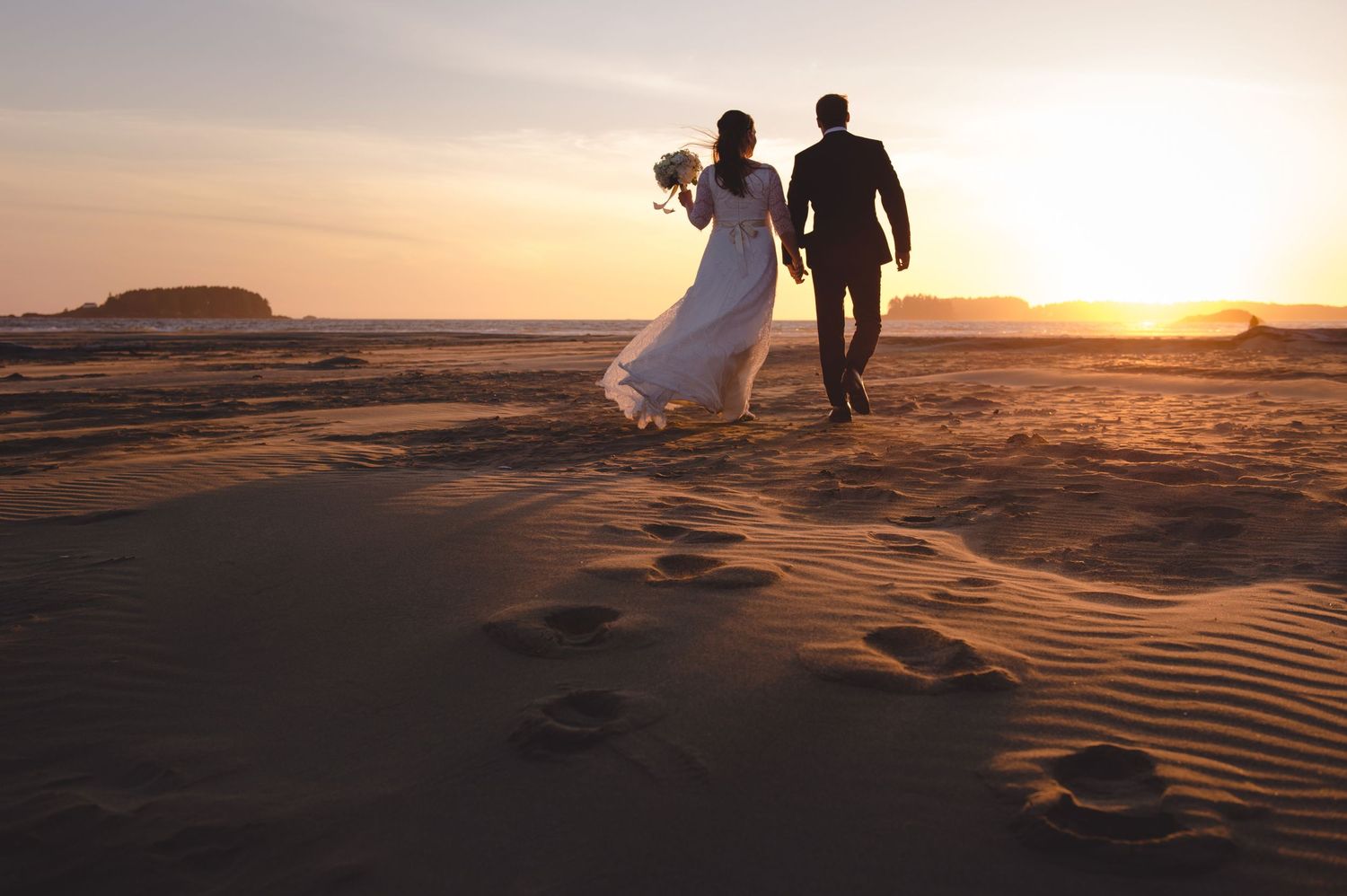 Wedding couple walks along sandy beach during golden hour with dramatic sunset lighting.