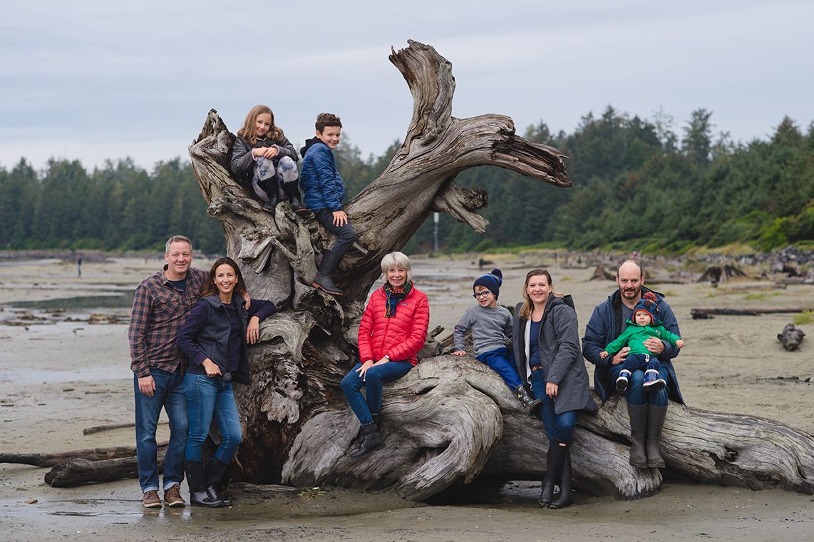 A group sits together on a large piece of driftwood on a scenic beach during overcast weather.