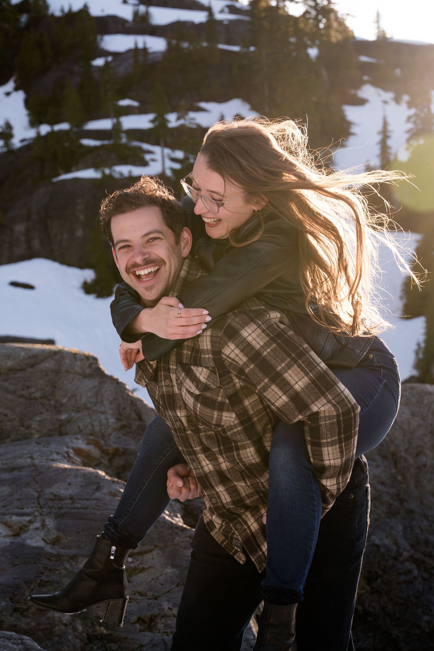 Joyful man giving woman a piggyback ride on rocky snowy mountain terrain at golden sunset.