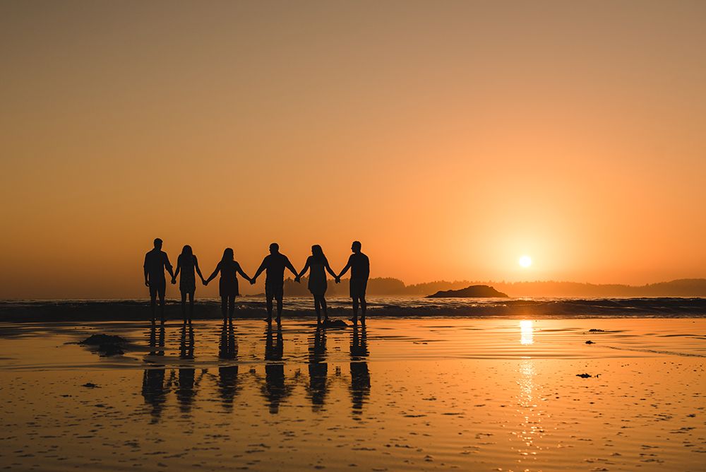 Silhouettes of people holding hands on beach at sunset with golden orange sky reflected in wet sand.