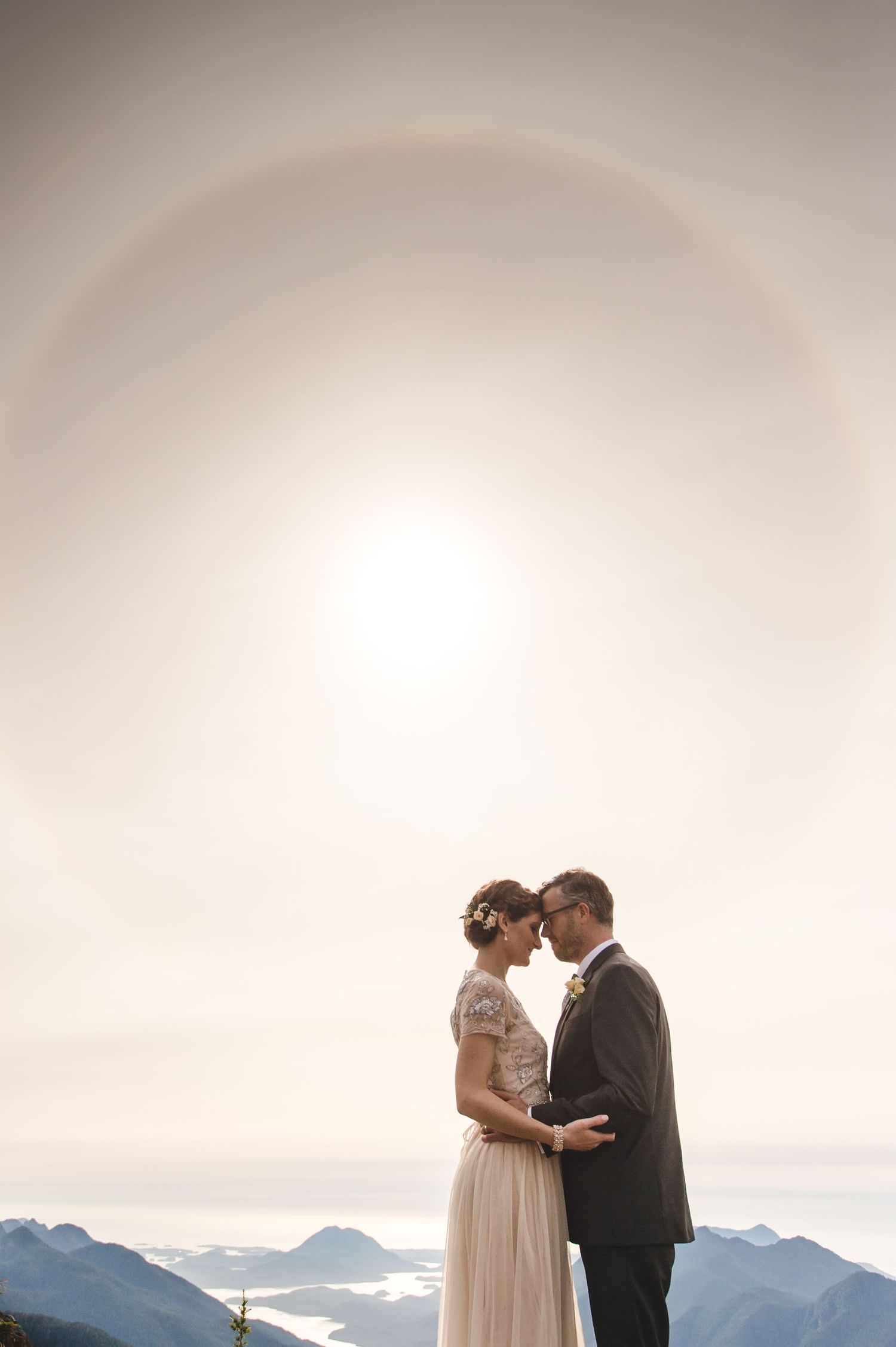 A romantic wedding couple embraces against a backdrop of misty mountain peaks at sunset.