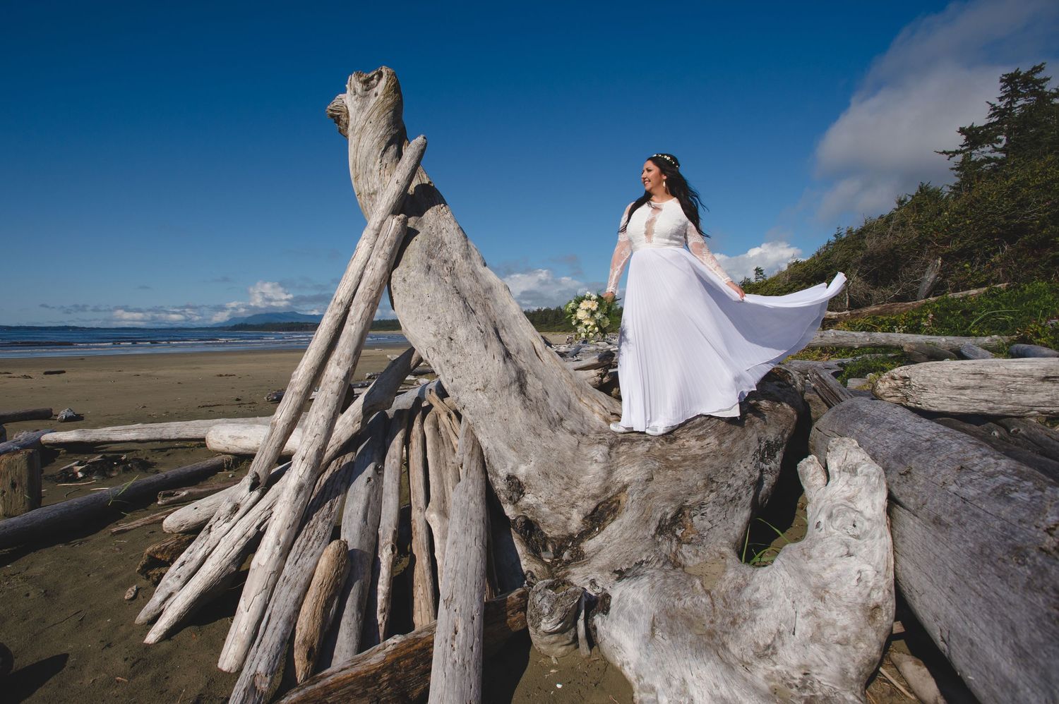 White wedding dress contrasts against weathered driftwood on a Pacific Northwest beach.