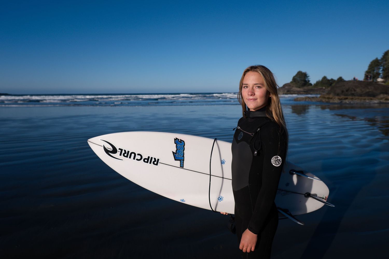 A surfer in a black wetsuit stands on a beach holding a white surfboard with blue ocean waves and coastline in background.