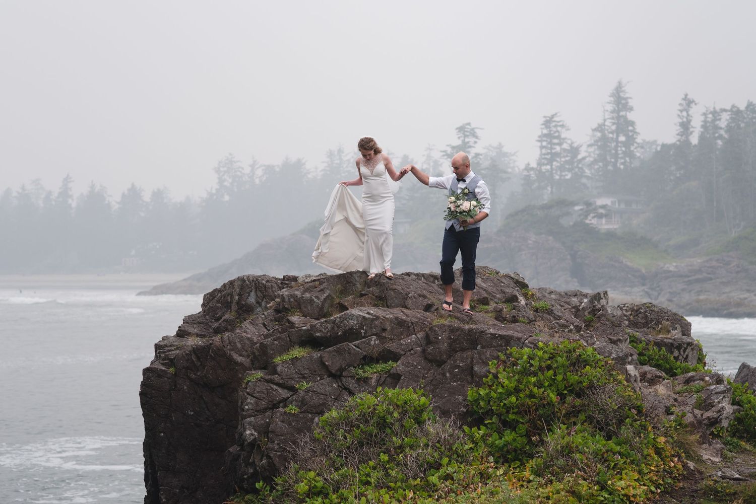 A couple celebrates joyfully on a rocky oceanside cliff during their wedding day.