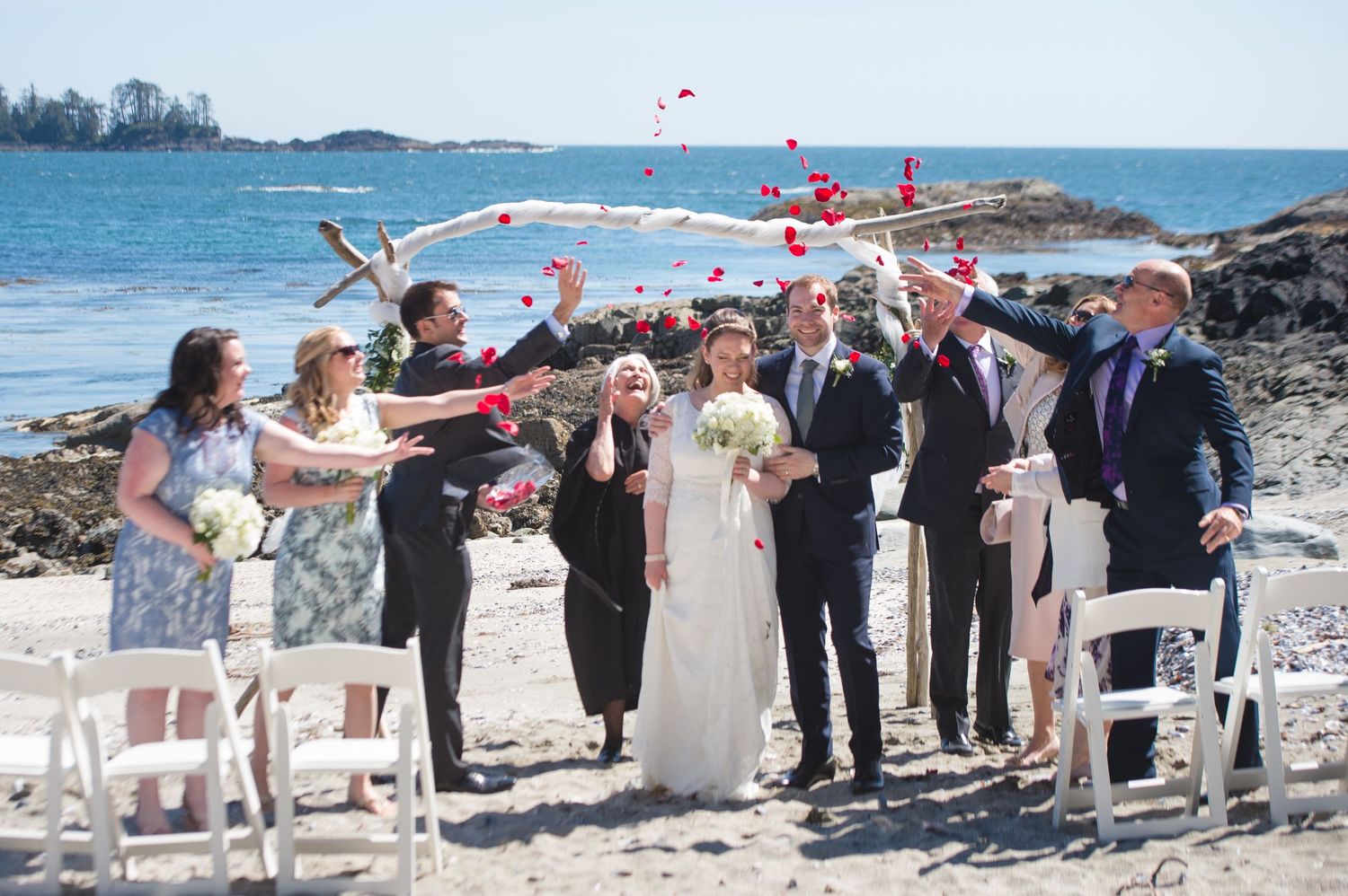 Wedding guests throwing flower petals during a beach celebration with ocean in background.