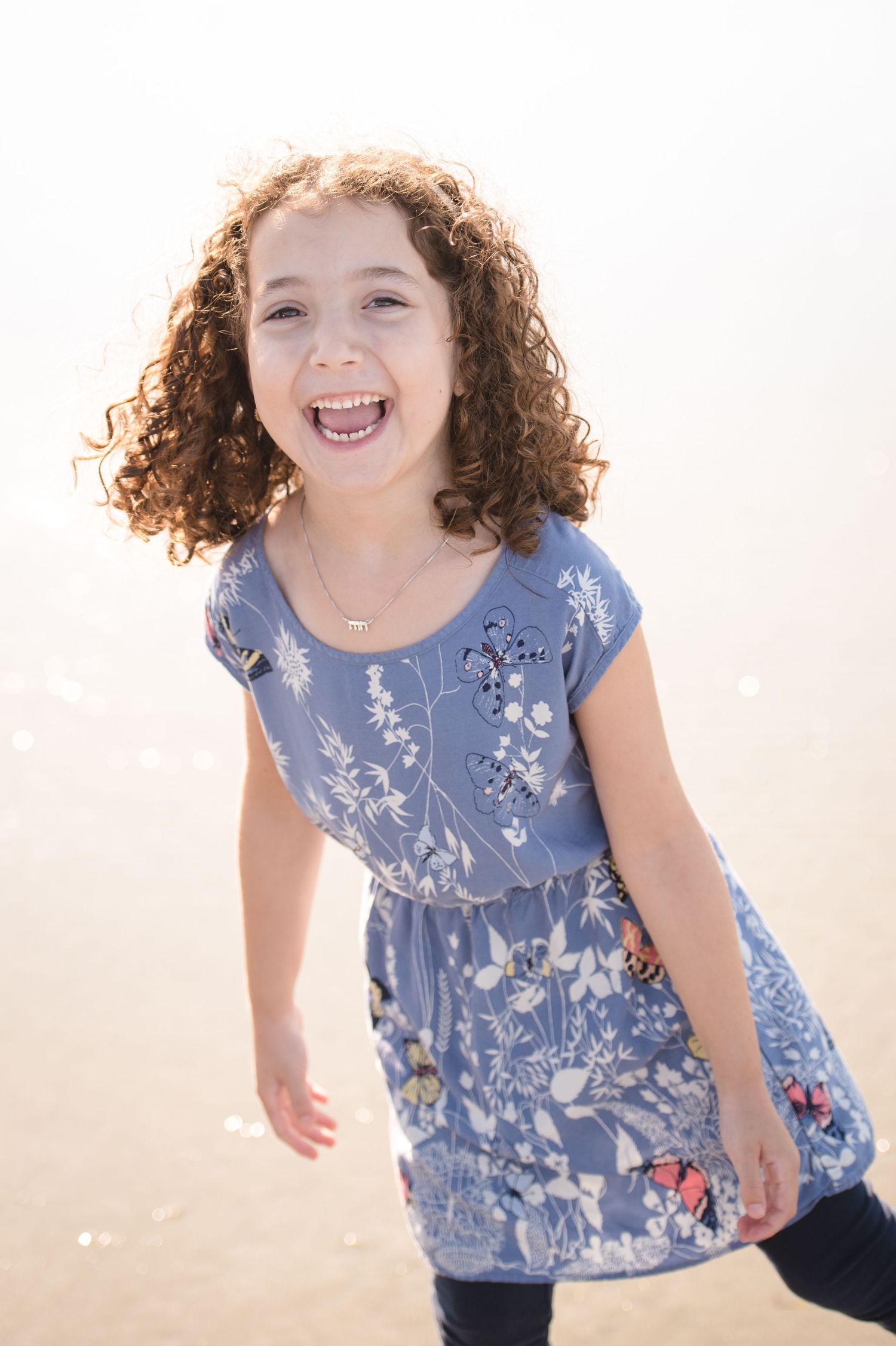 A joyful child wearing a blue floral dress laughs while standing against a bright background.