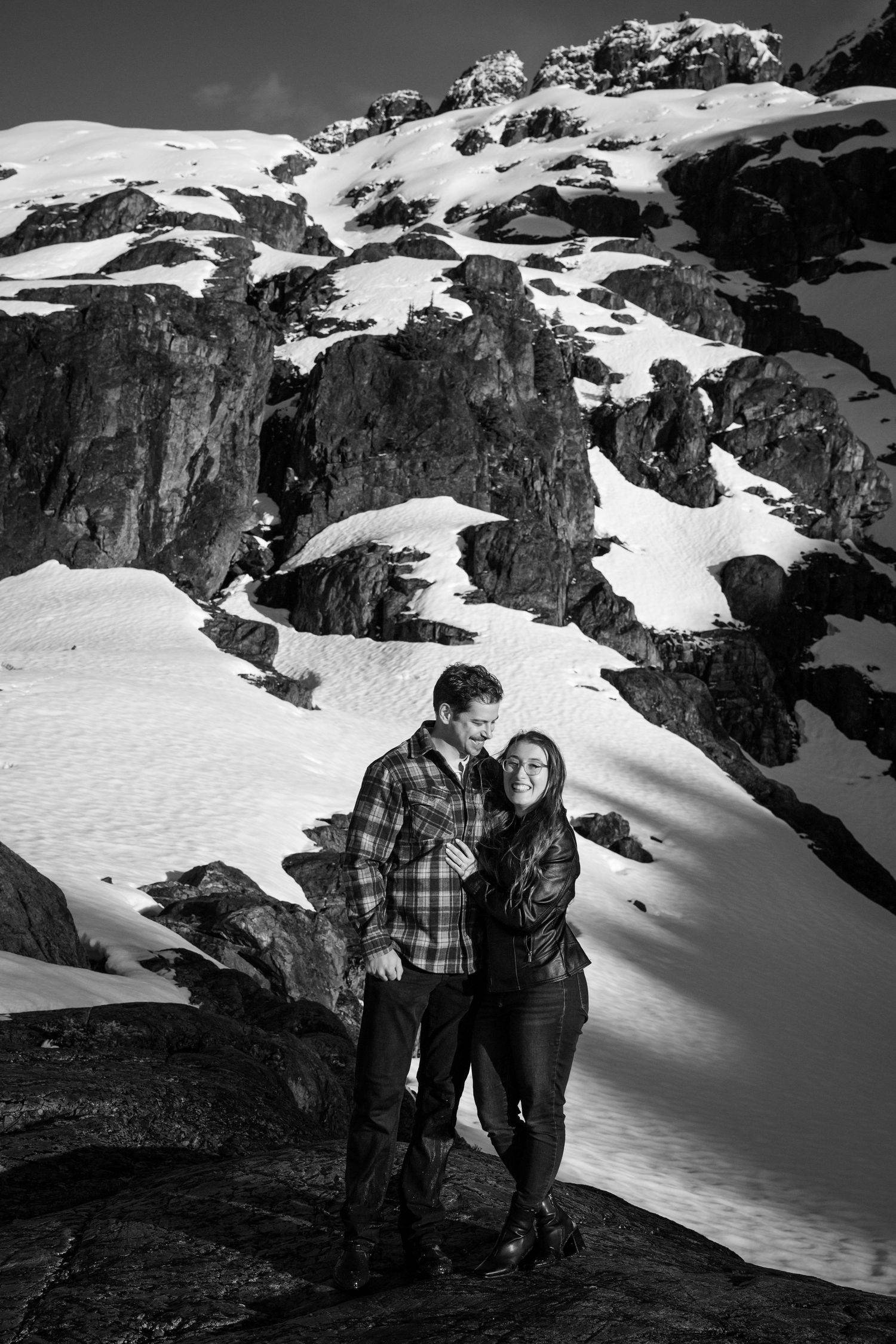 Black and white photo of a couple embracing on a snowy mountain rocky landscape during winter.