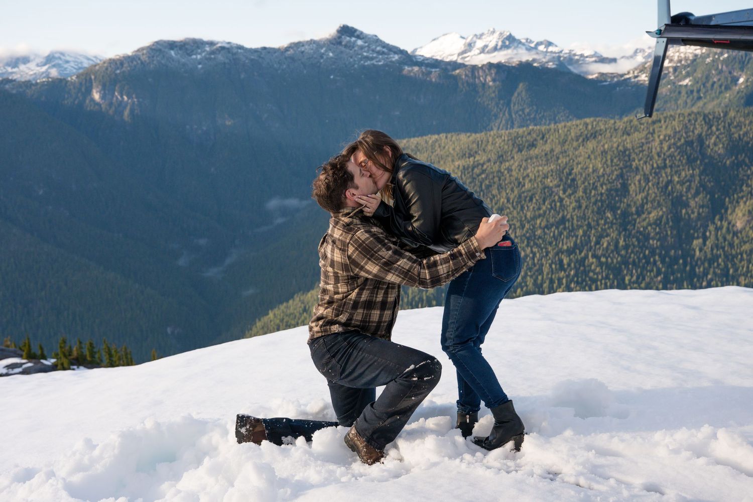 Couple sharing a romantic marriage proposal moment on a snowy mountain peak with scenic valley views.