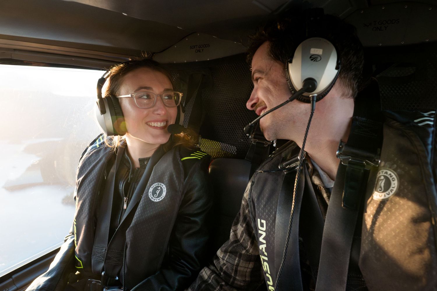 Two people wearing headsets smile at each other inside a helicopter cockpit during a scenic flight.
