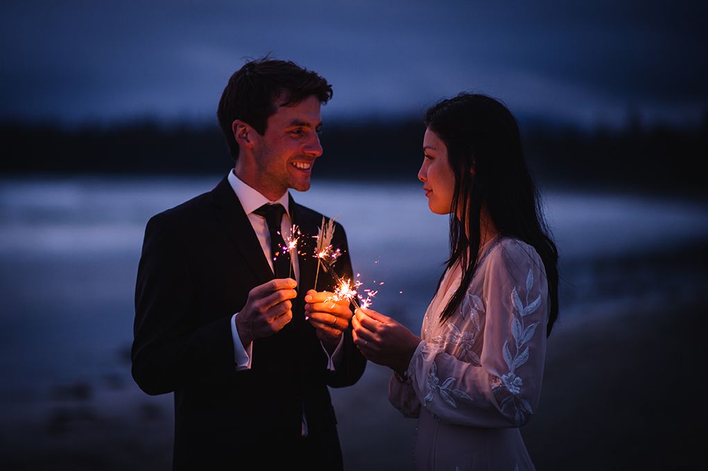 Wedding couple shares an intimate moment with sparklers at twilight by the ocean.