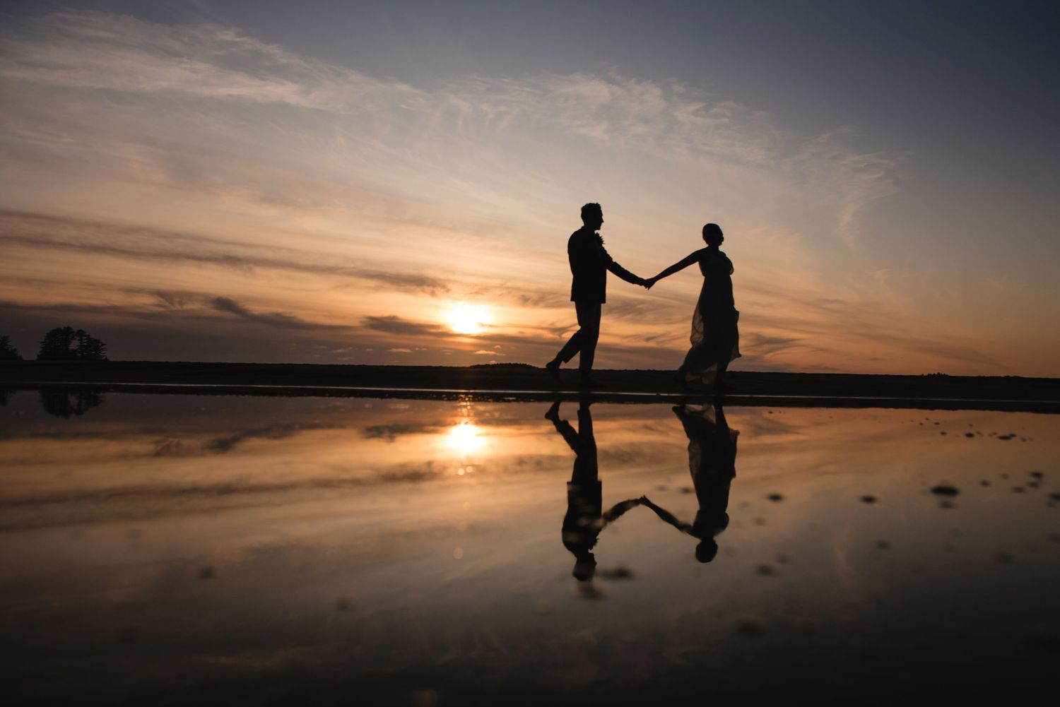 A silhouetted couple holds hands against a dramatic sunset sky reflected in wet beach sand.