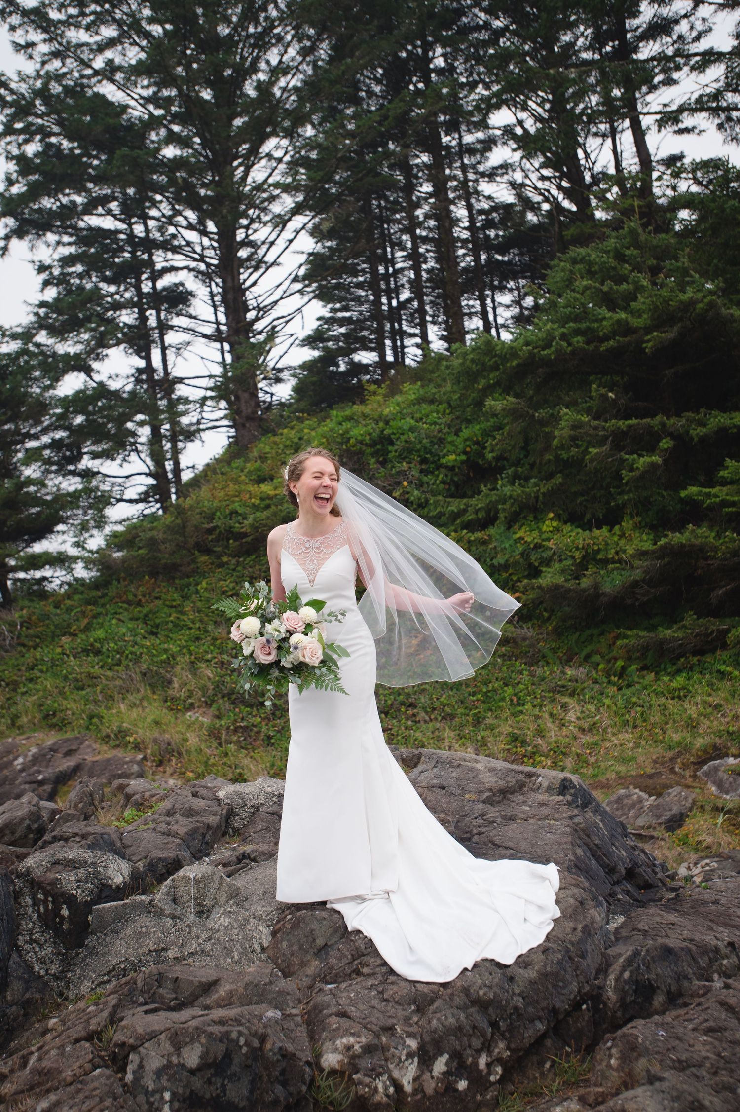A bride in a white wedding dress poses on rocky coastal cliffs with evergreen trees in the background.