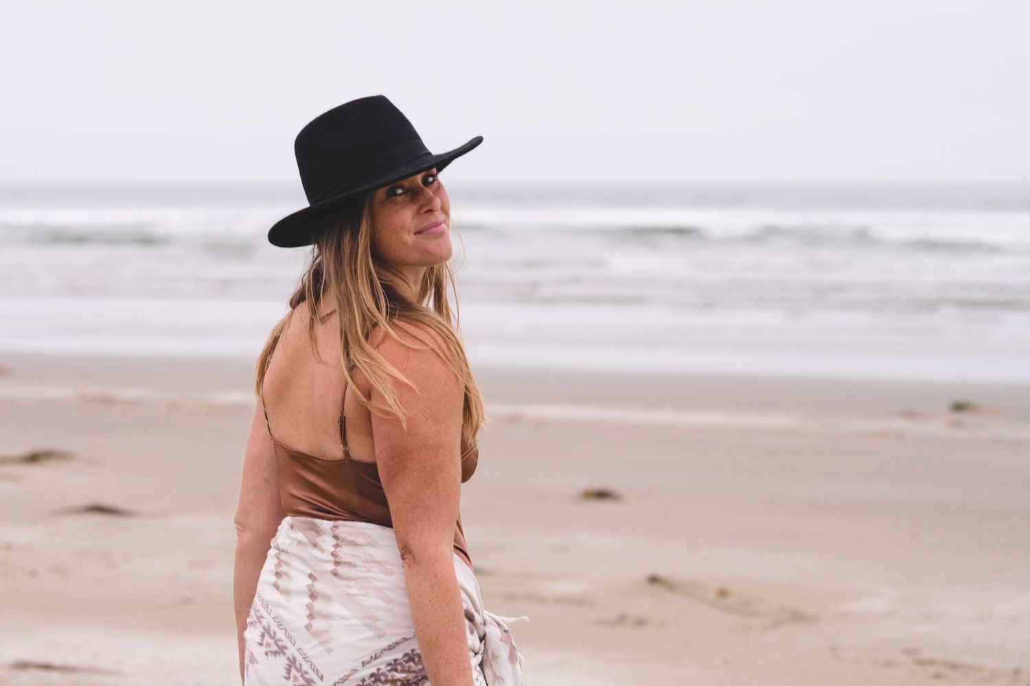 A person in a black hat walks along a misty beach shoreline while waves crash in the background.