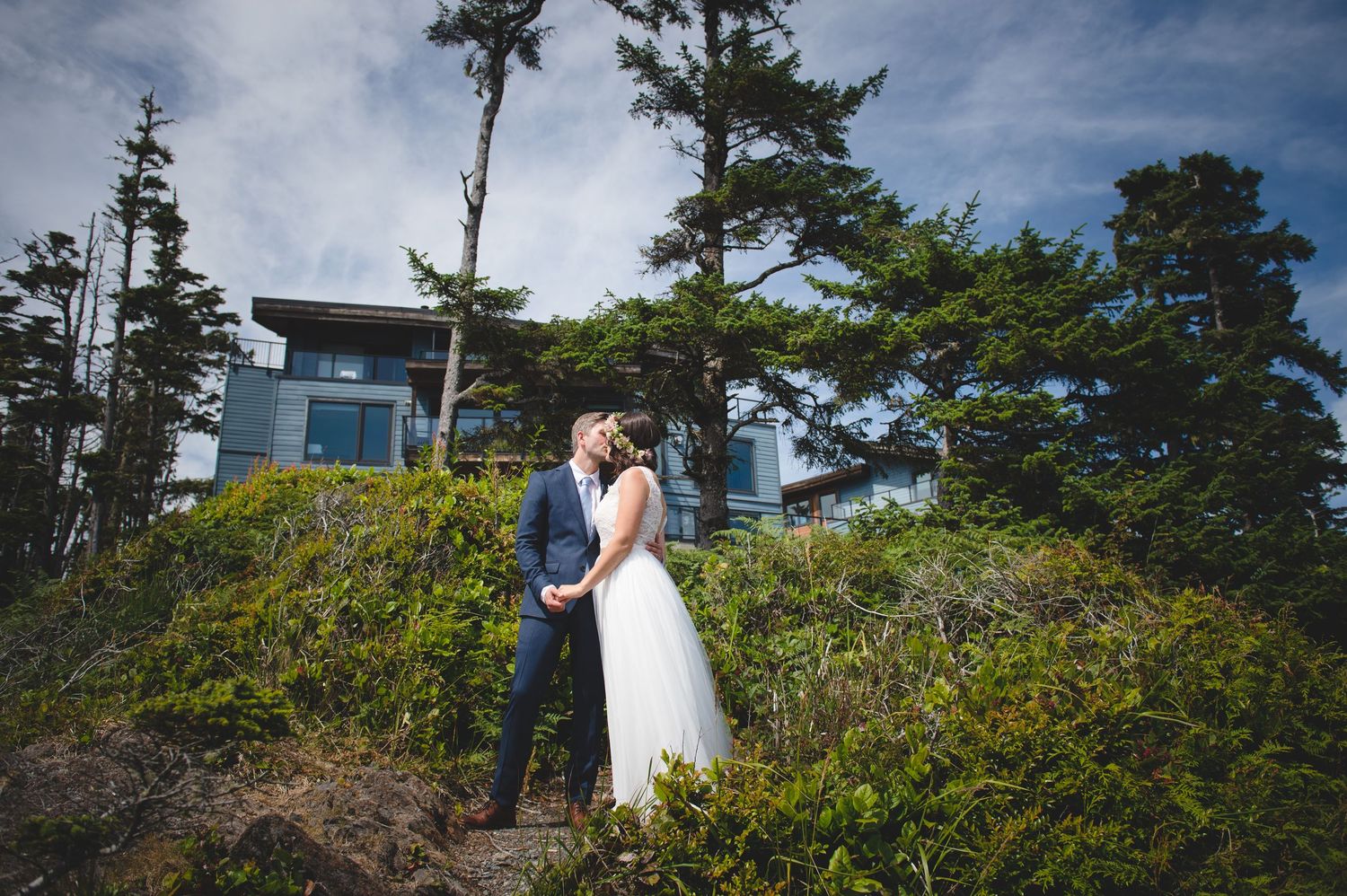 A couple shares an intimate moment on a coastal hillside with a modern house in the background.