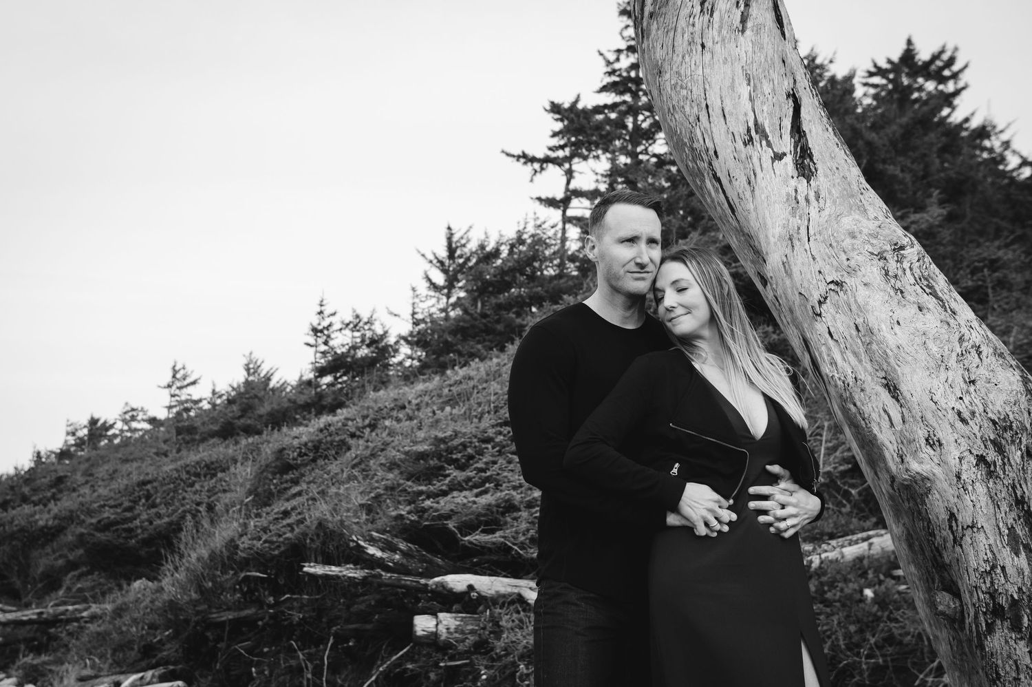Couple in dark clothing embracing near trees on cloudy day.