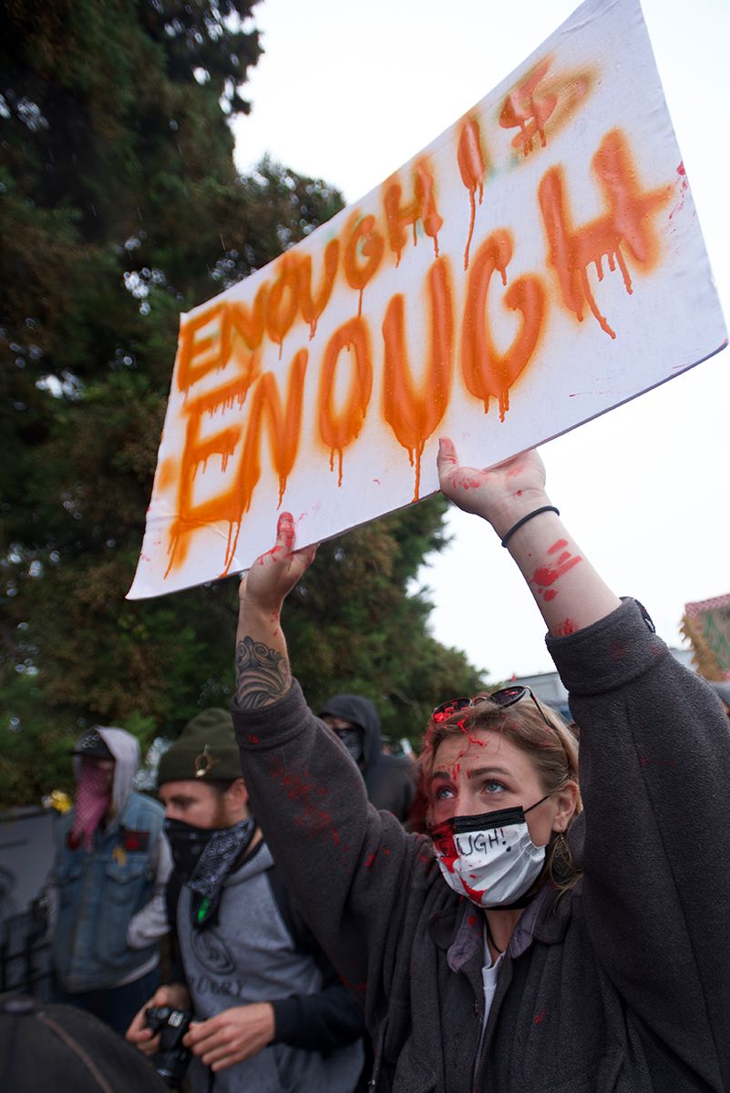 Someone holding up a protest sign with orange text against an overcast sky.