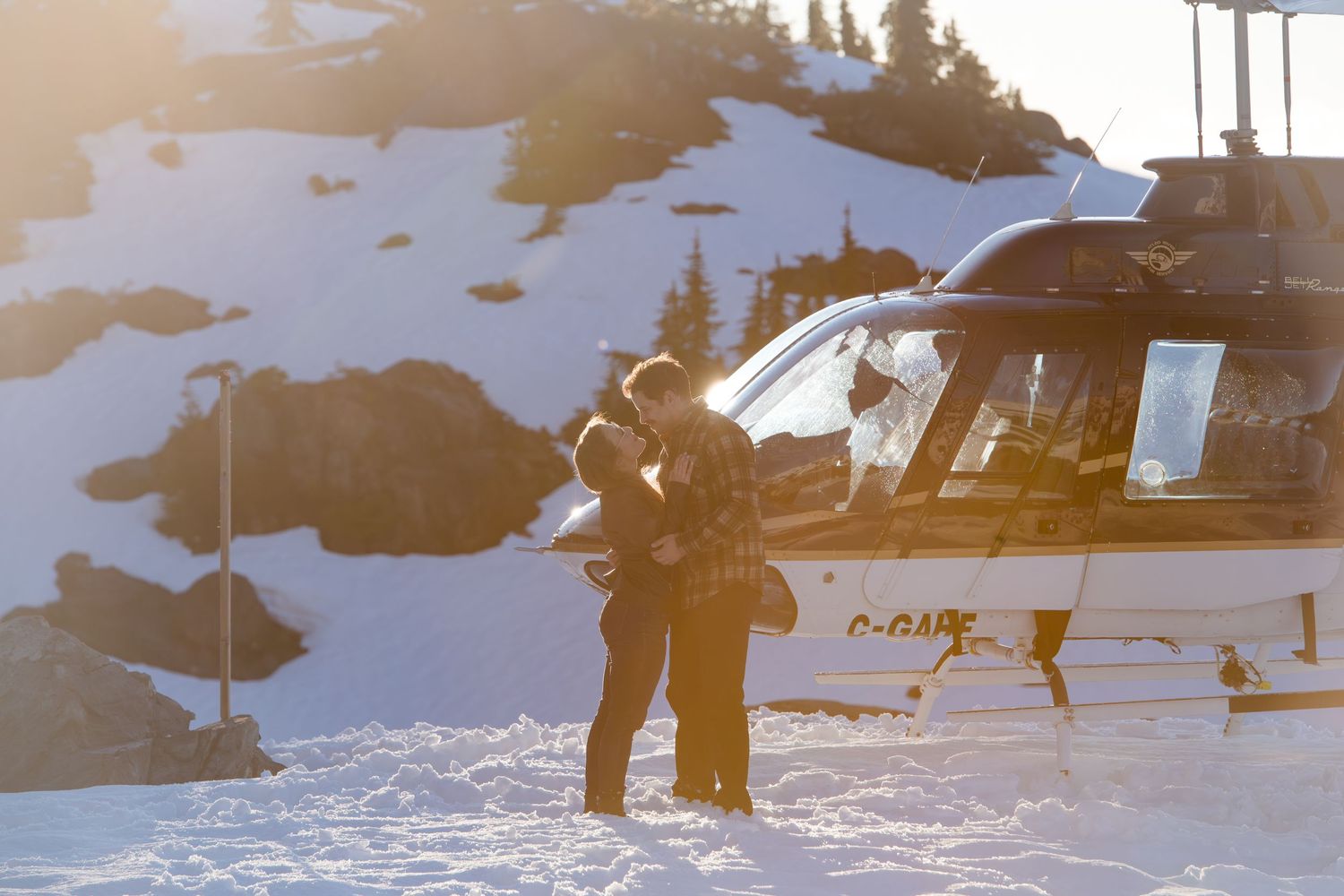 Two people stand beside a helicopter on a snowy mountain landscape bathed in golden winter sunlight.