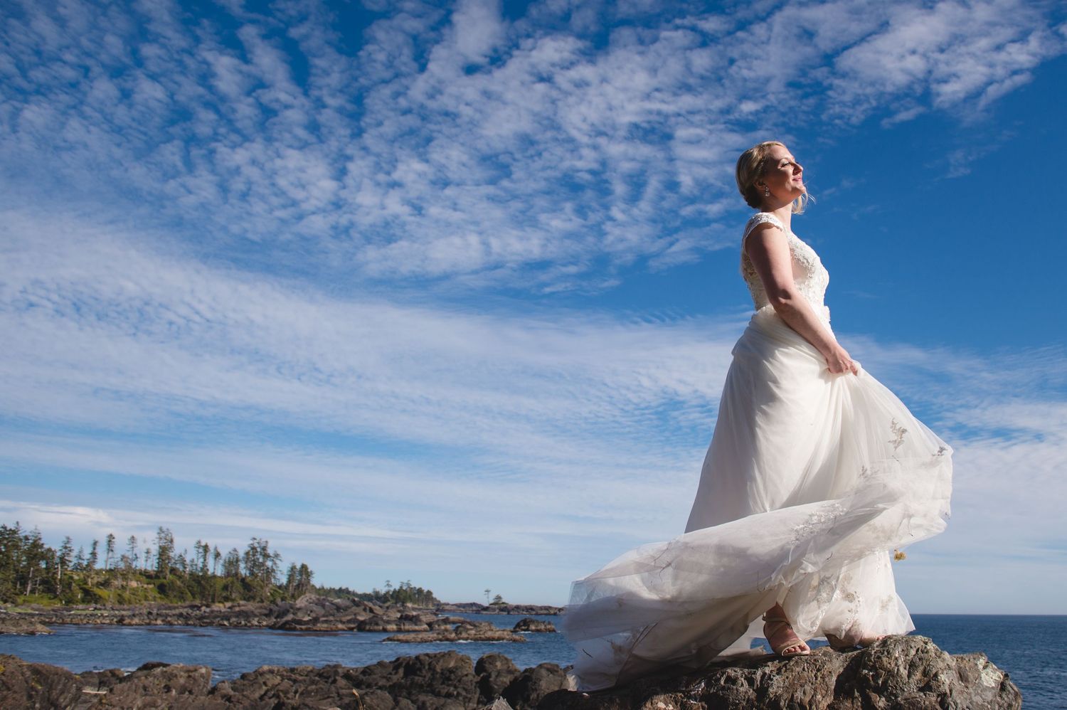 Bride in flowing white dress twirls against dramatic coastal landscape and blue skies.