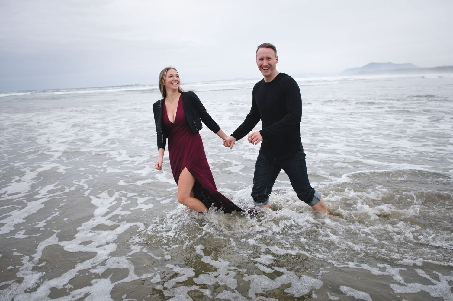 A couple wearing black and burgundy outfits plays in the ocean waves on an overcast day.
