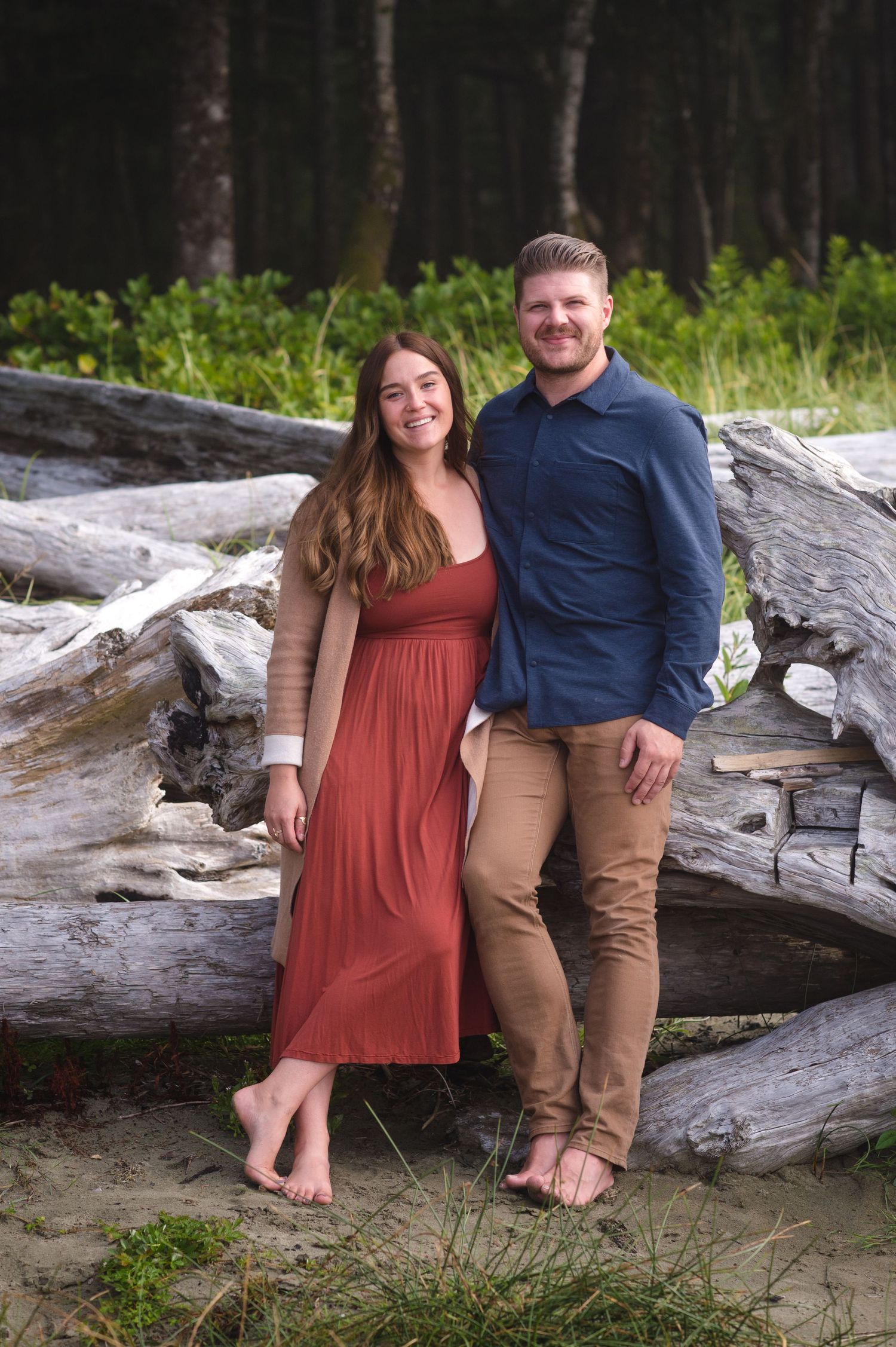 A couple in casual attire stands together on driftwood at a beach near evergreen trees.