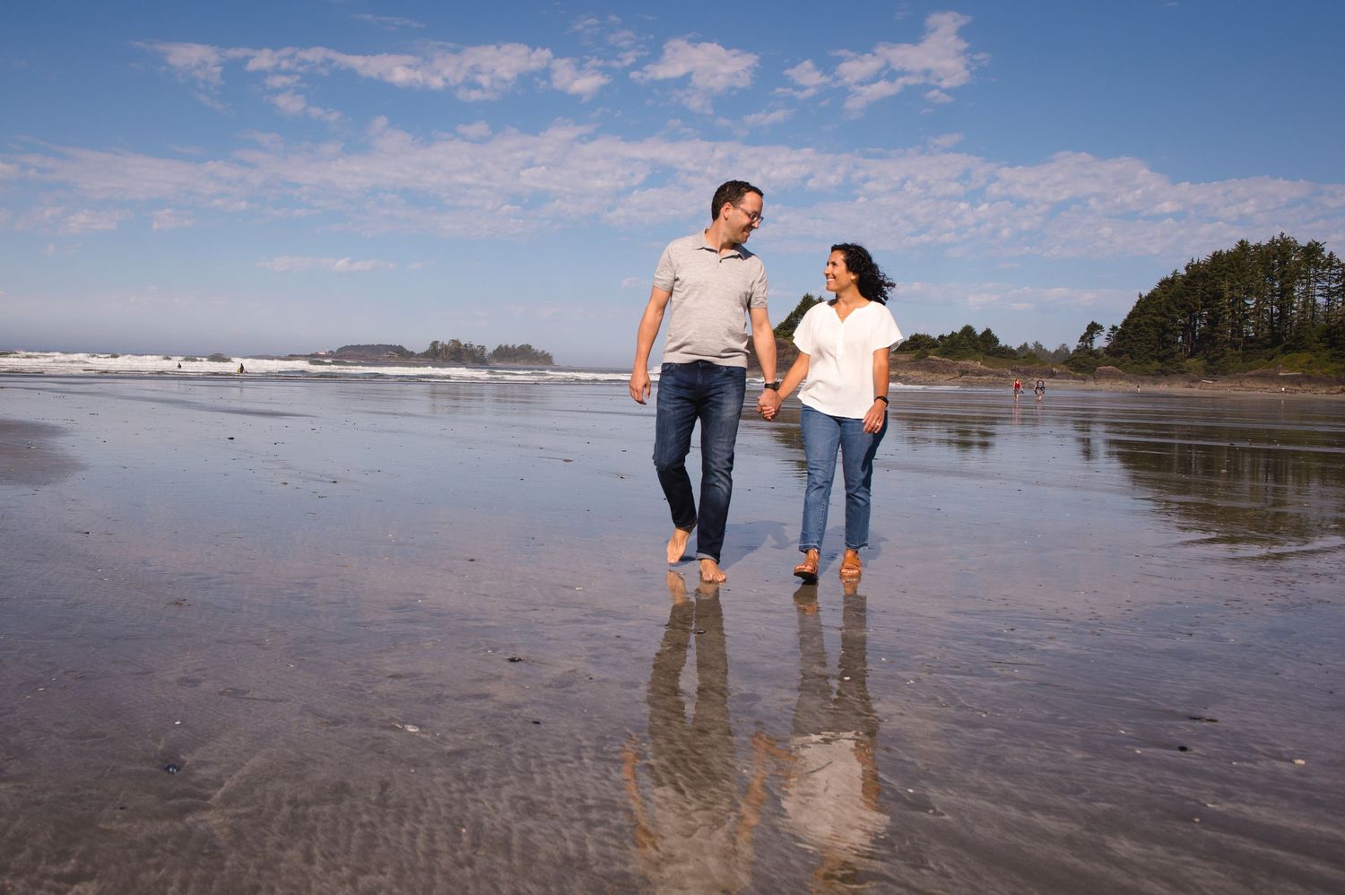 A couple walking hand in hand on a wet beach with waves and mountains in the background.