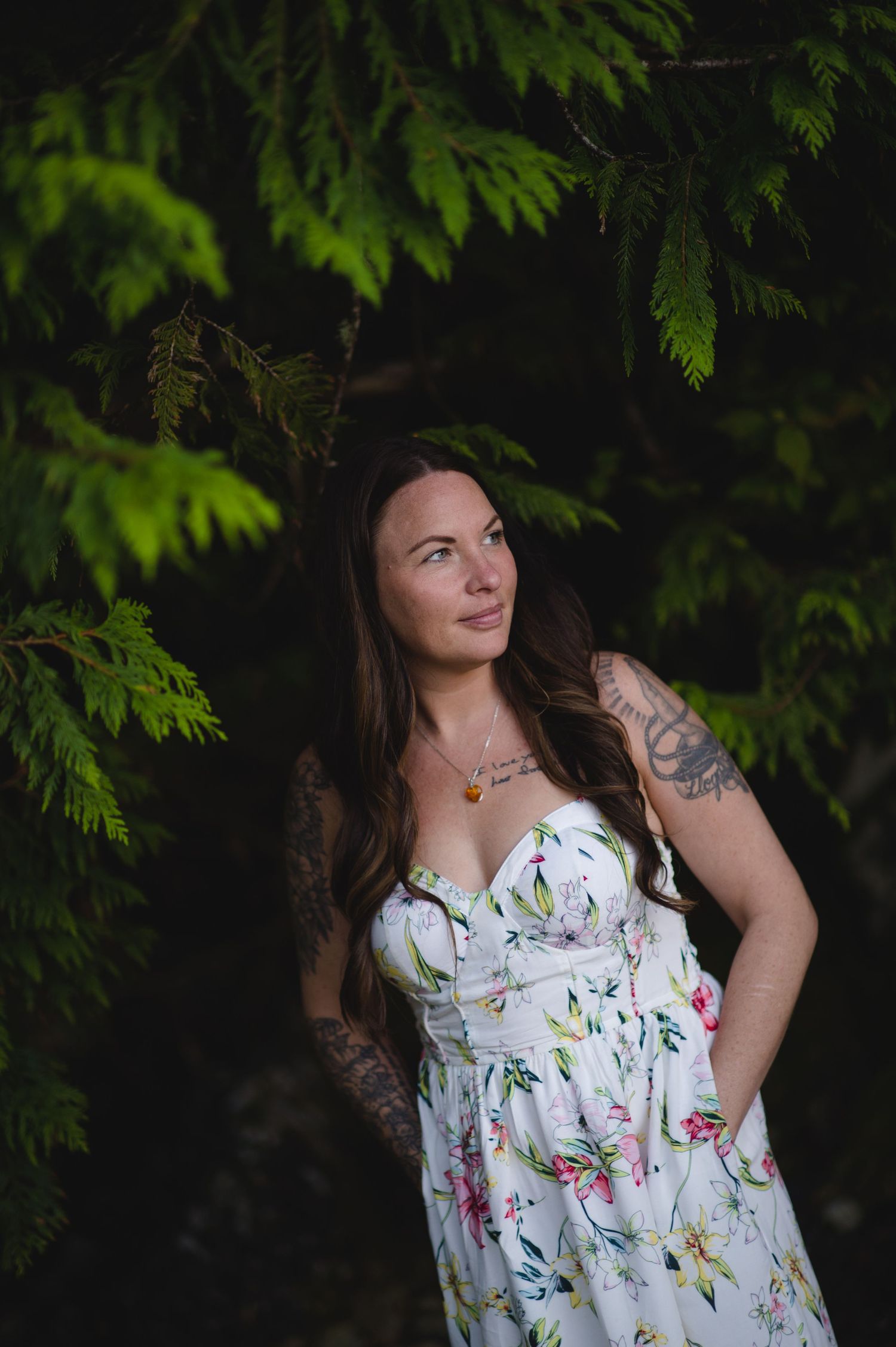 A woman in a sleeveless floral dress stands in front of lush green foliage.
