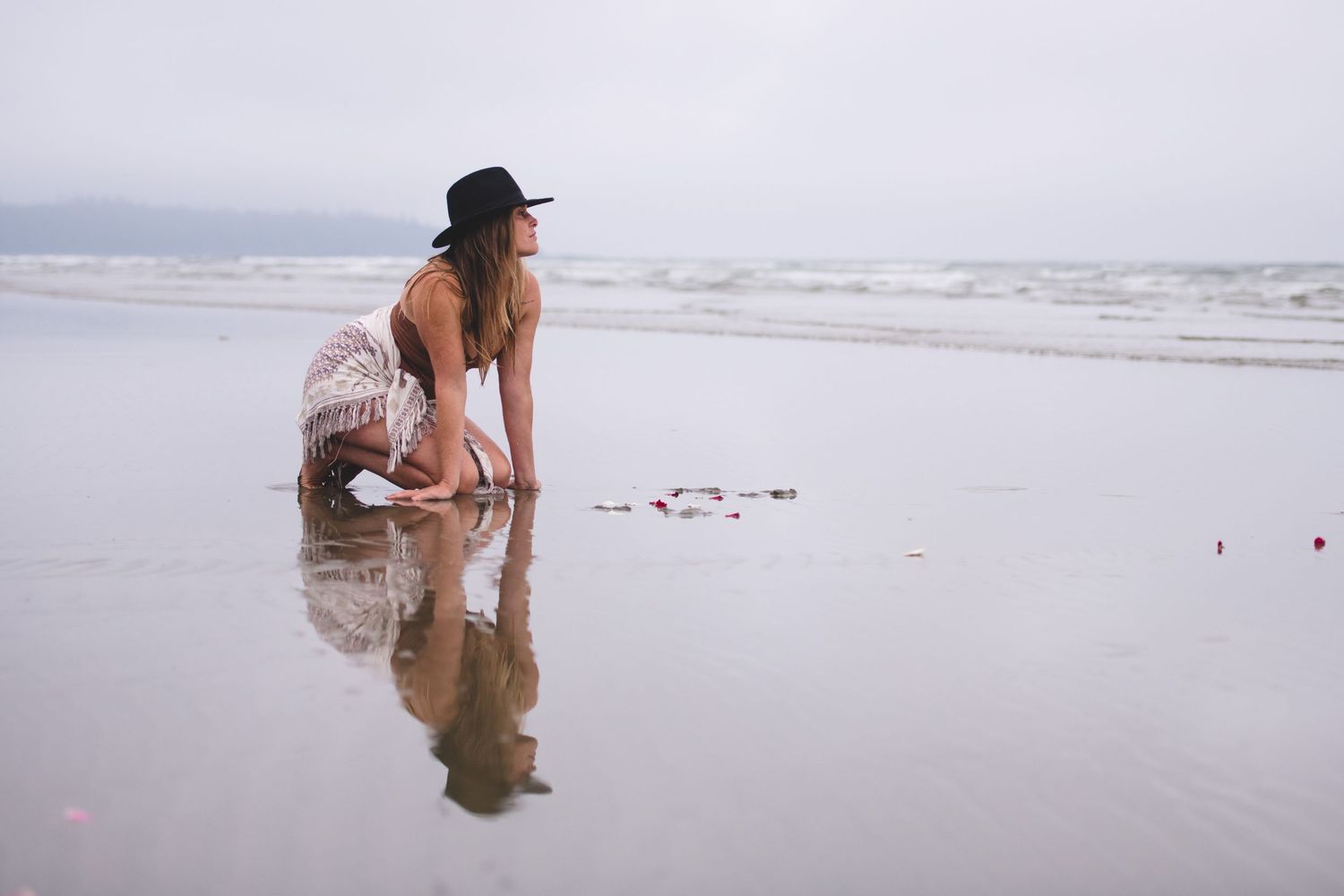 Figure in hat explores shallow ocean waters on overcast beach day.