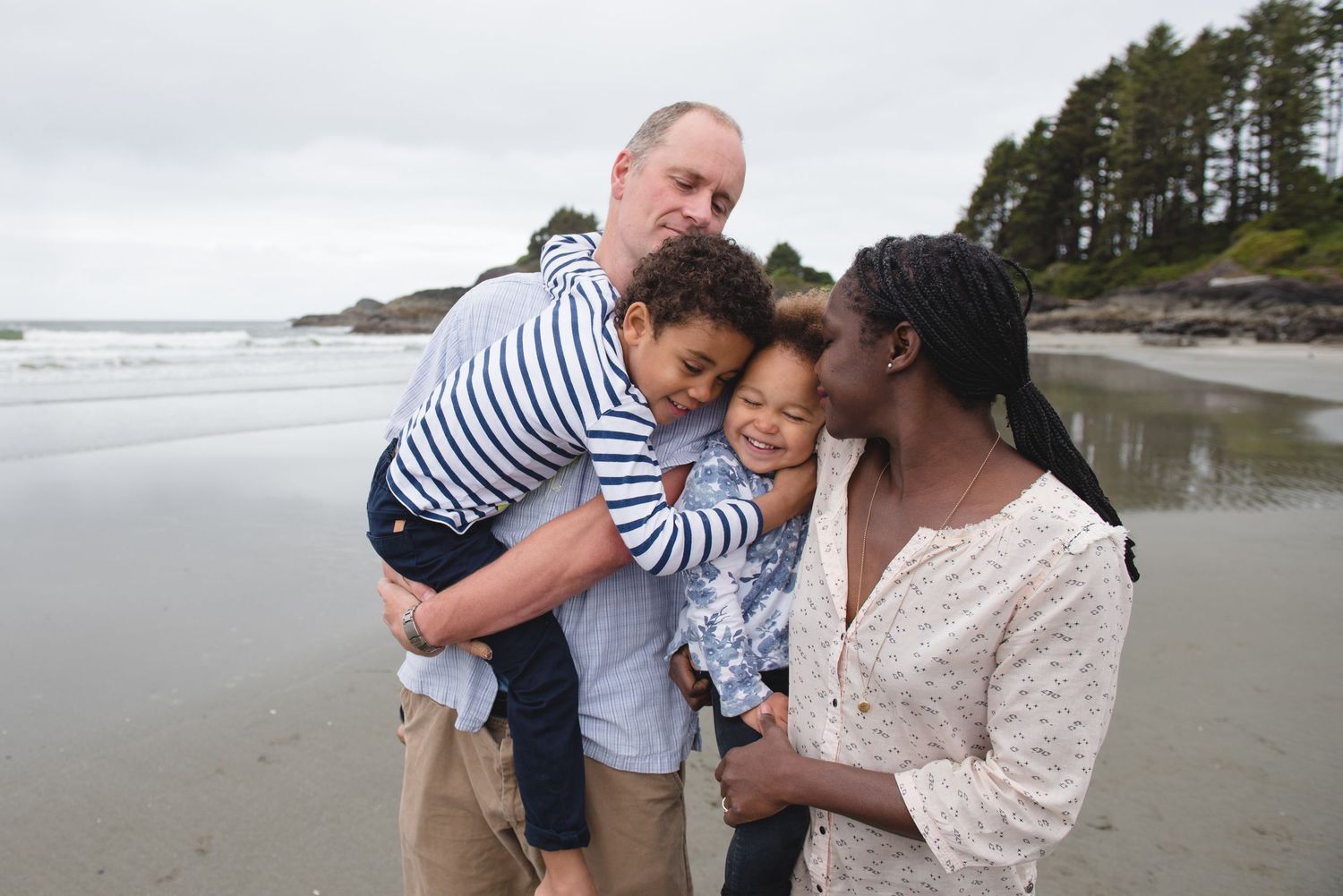 Family enjoying a playful moment together on a scenic beach with evergreen trees and coastline in the background.