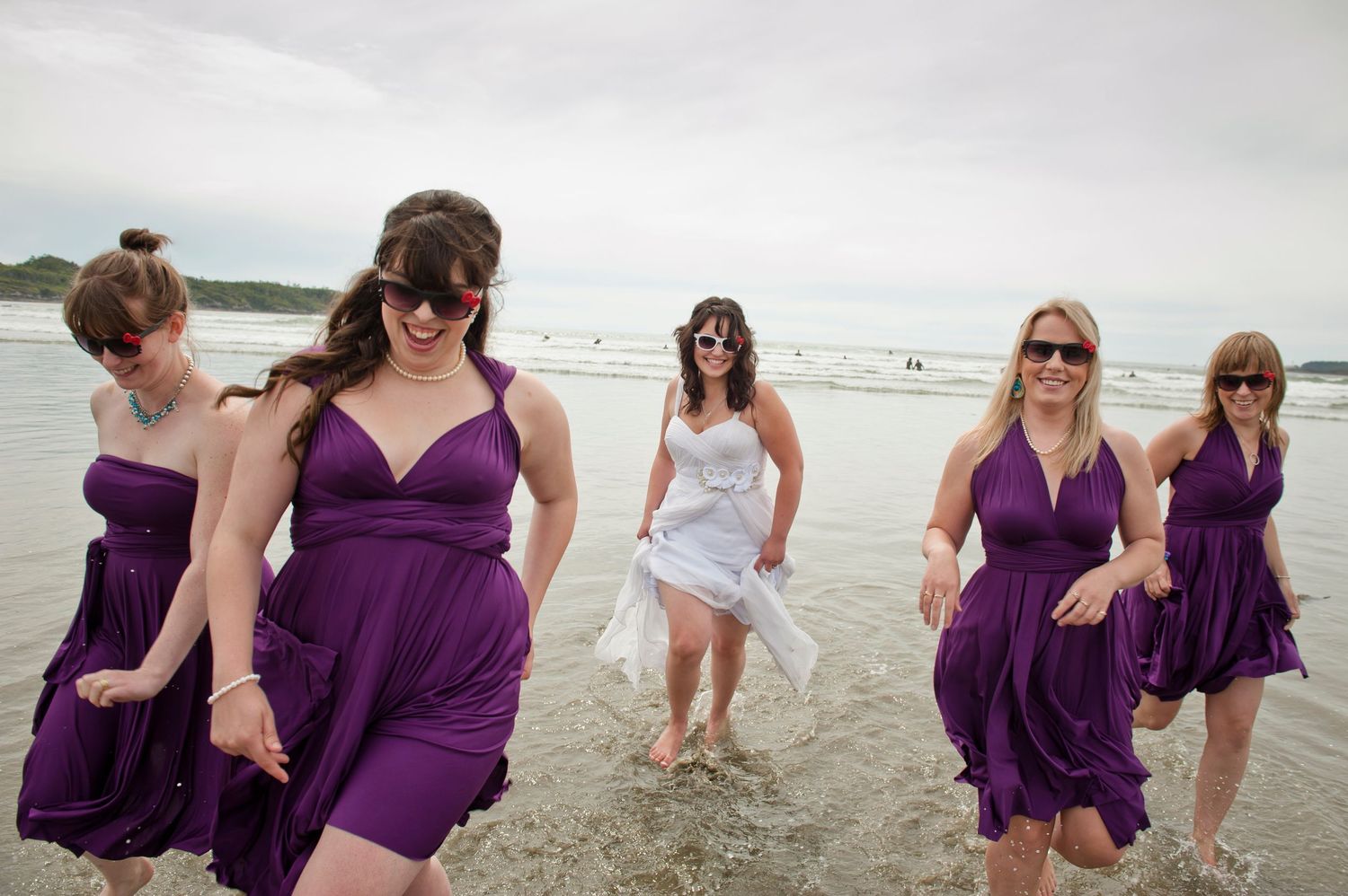 Bridesmaids in purple dresses playing and laughing on a beach.
