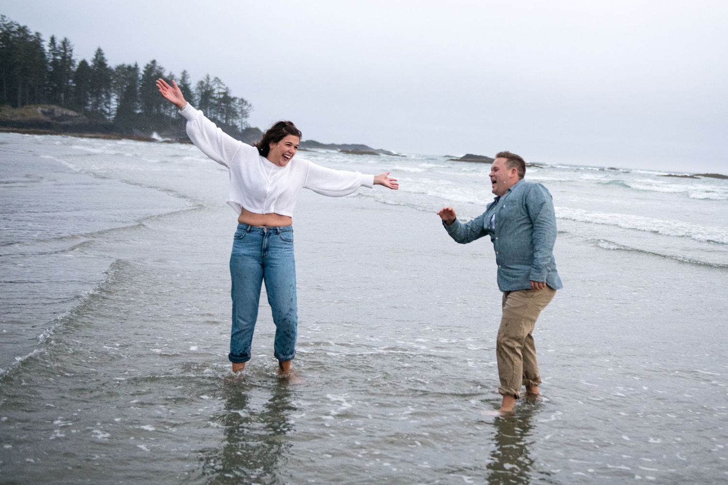 Couple holds hands while wading through ocean waves on a cloudy beach day.