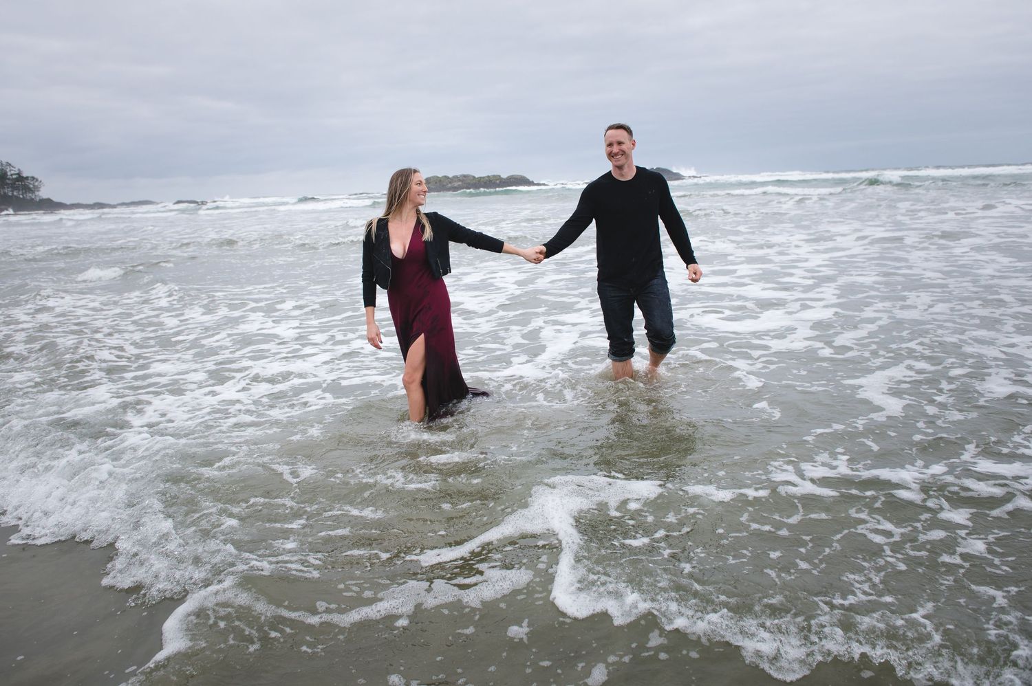 A couple wades through shallow ocean water while holding hands on a cloudy Pacific Northwest beach.