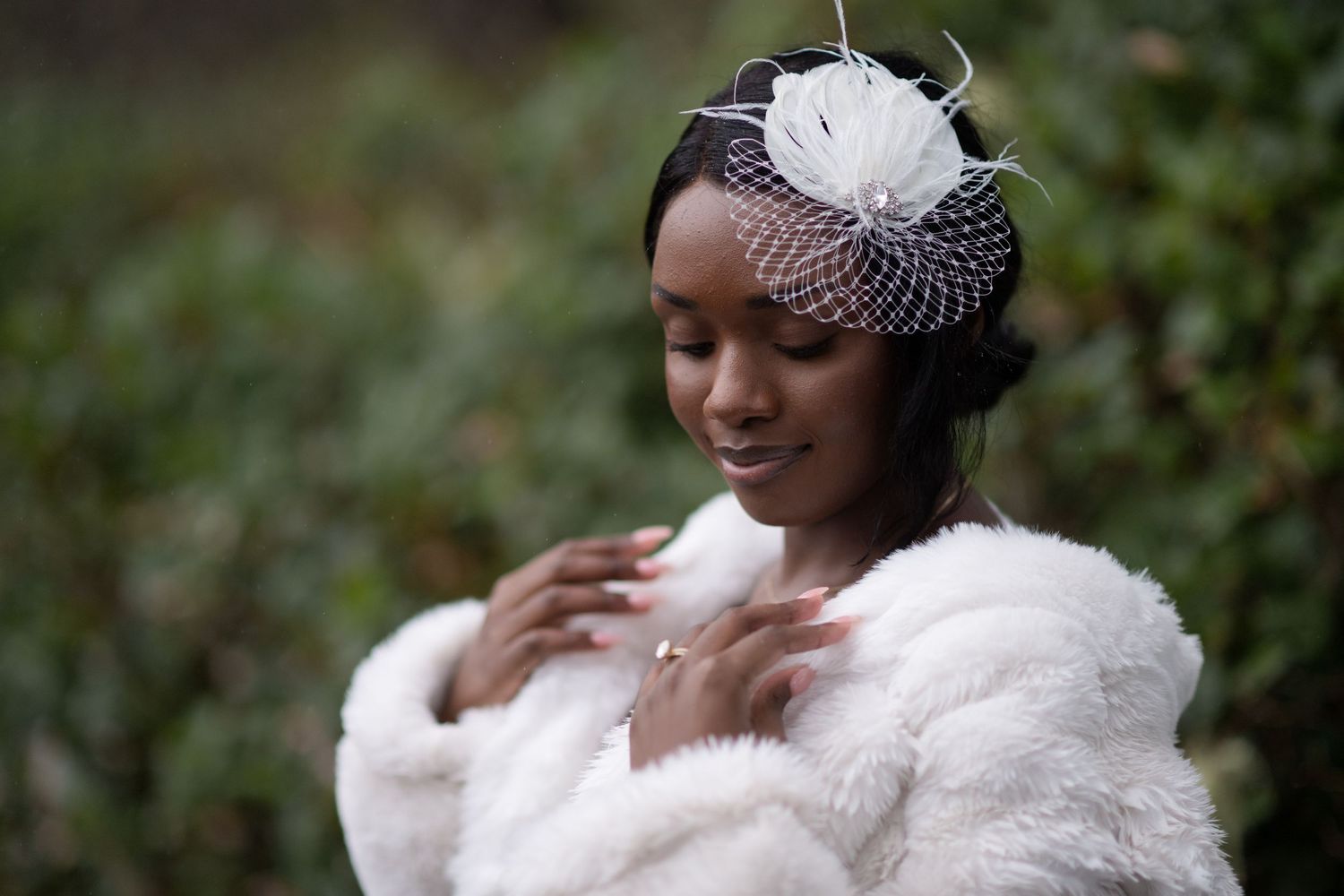 A bride wearing a white fur coat and vintage-style fascinator poses elegantly in a garden setting.