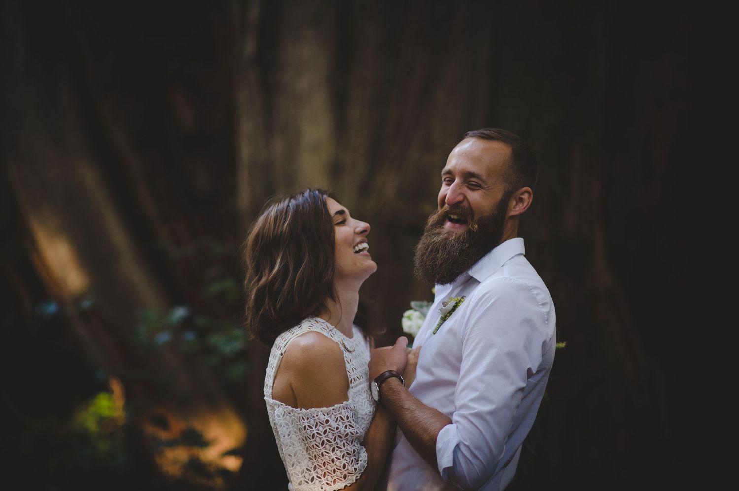 A couple shares an intimate moment while laughing together in a wooded setting with filtered sunlight.