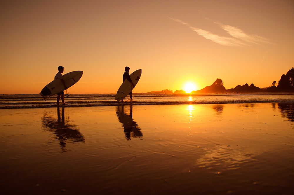 Surfers standing on beach at golden sunset with reflections on wet sand.