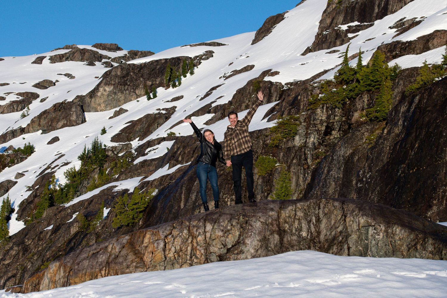 Couple standing on a rocky ledge surrounded by snow-covered mountain cliffs under blue sky.