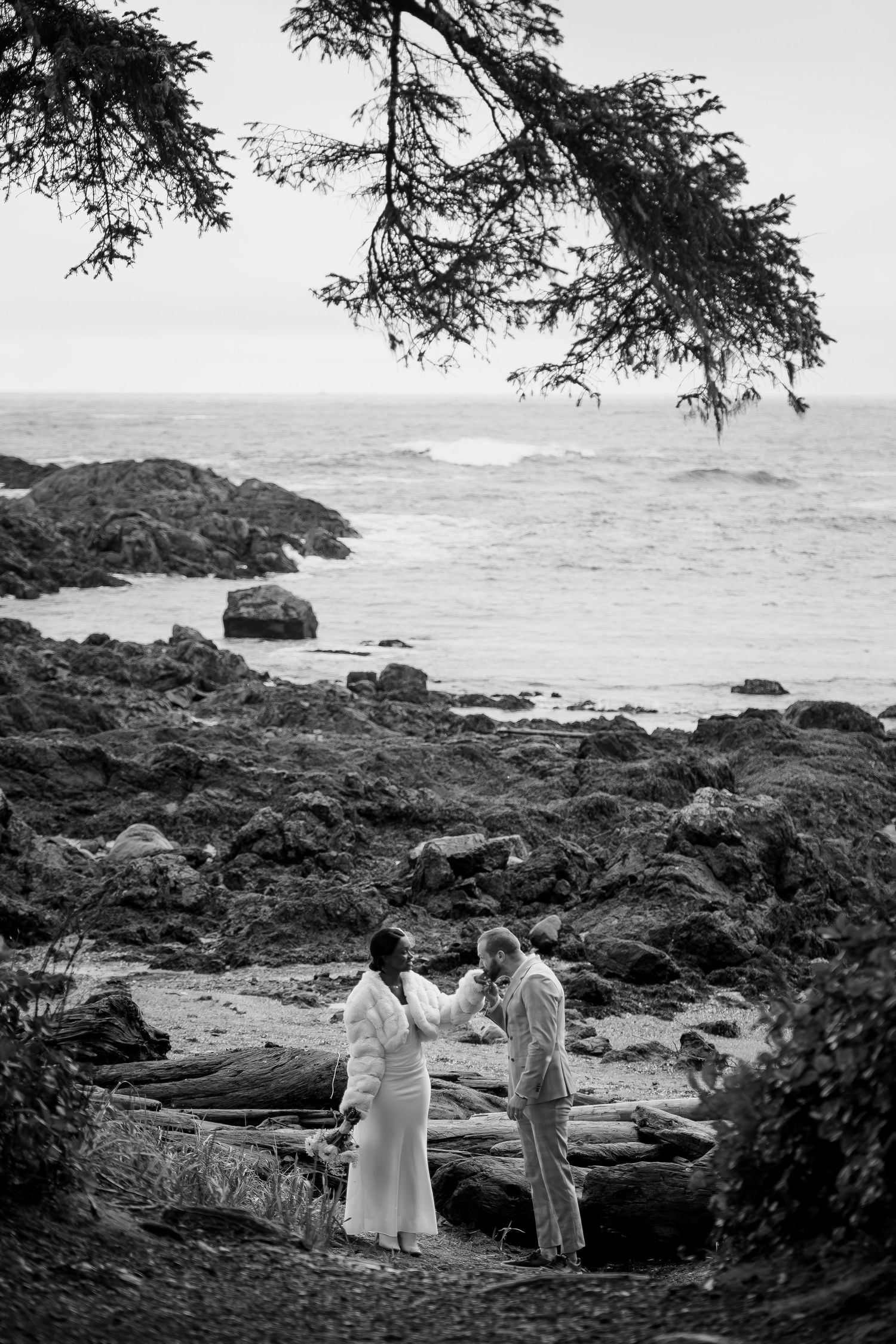 A black and white wedding ceremony photo framed by nature on a dramatic rocky coastline.