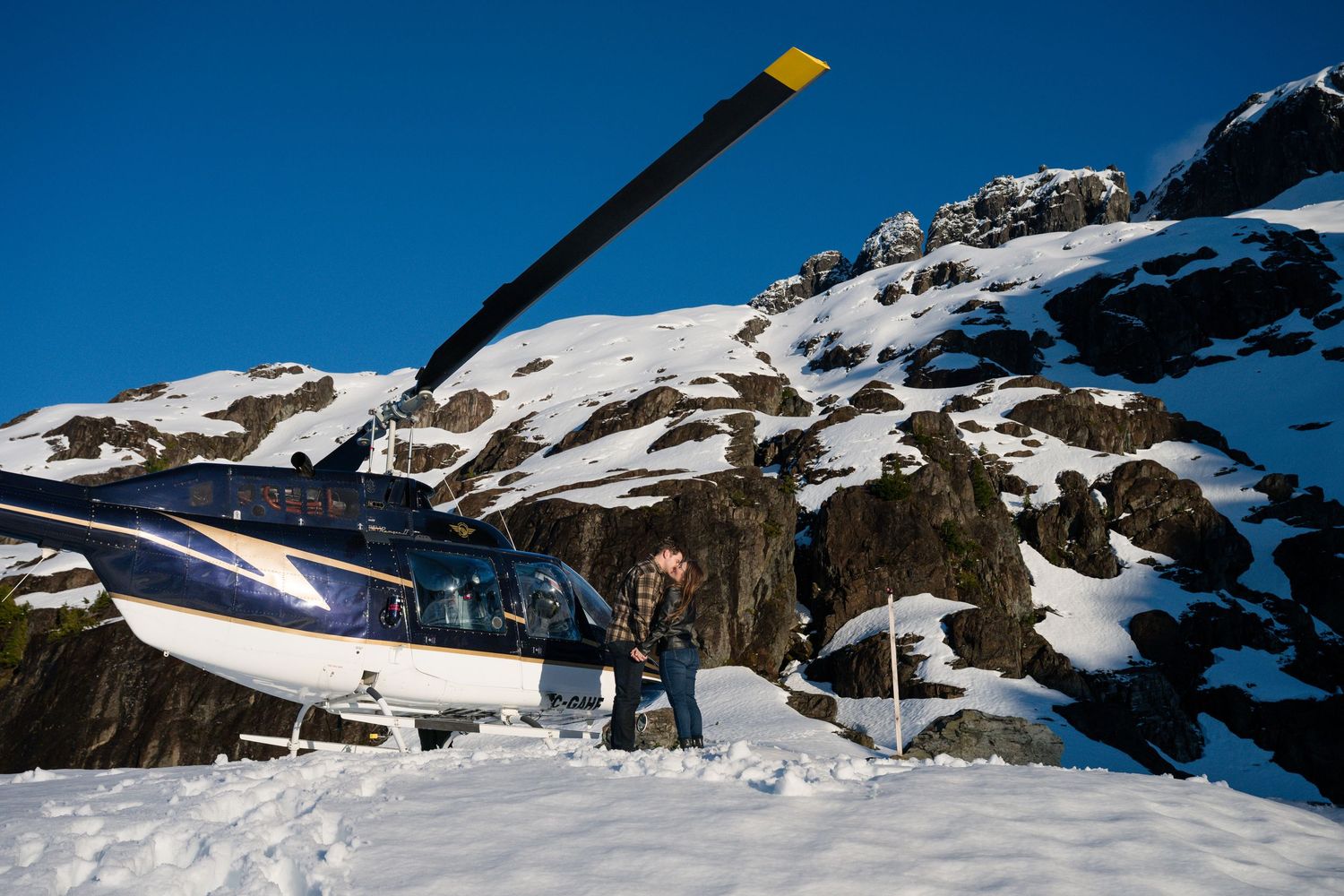 Couple standing together beside a helicopter on a remote snowy mountain peak surrounded by rocky terrain.