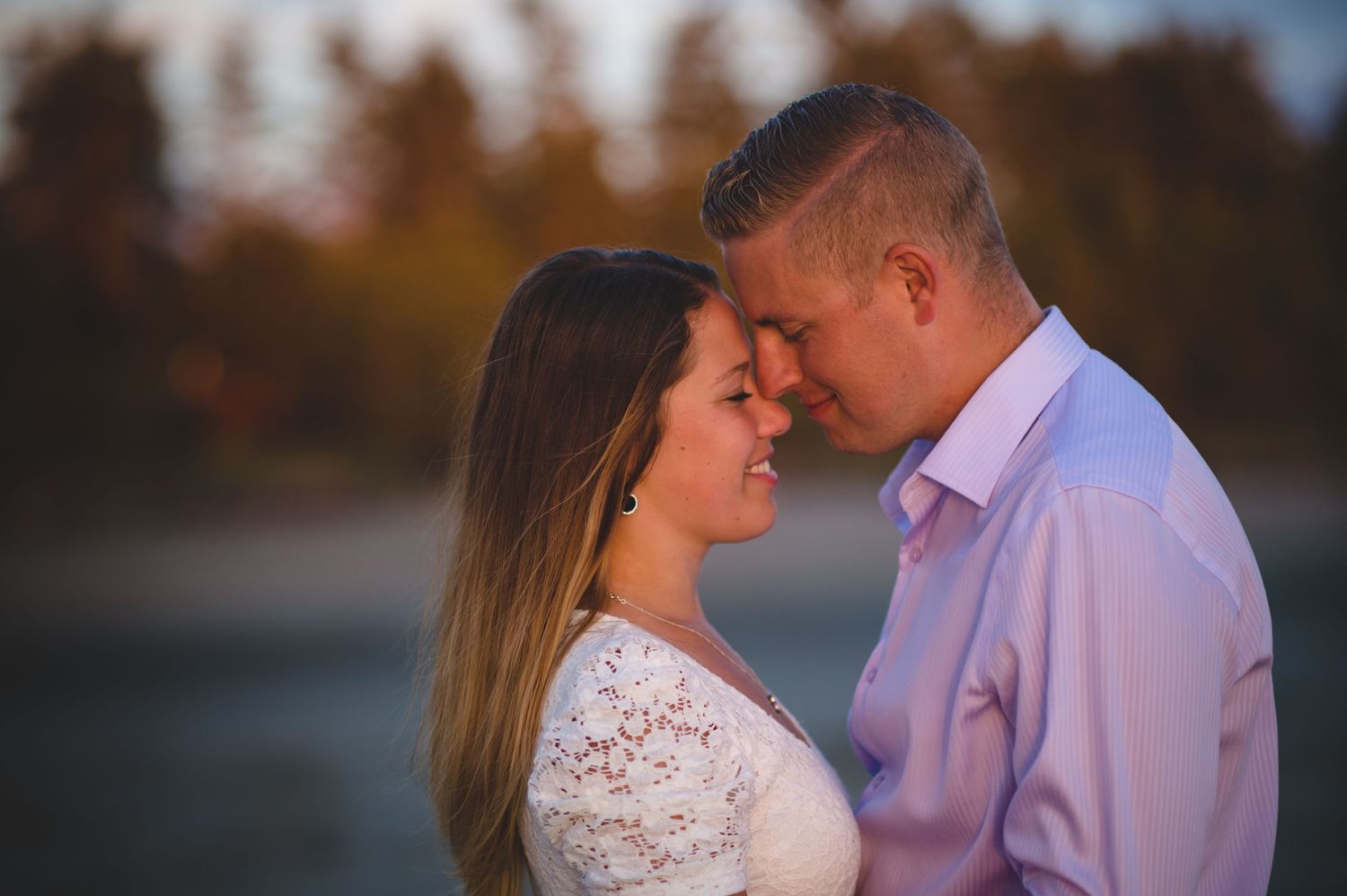 A romantic couple in formal attire embraces during an autumn sunset engagement photoshoot by a lake.