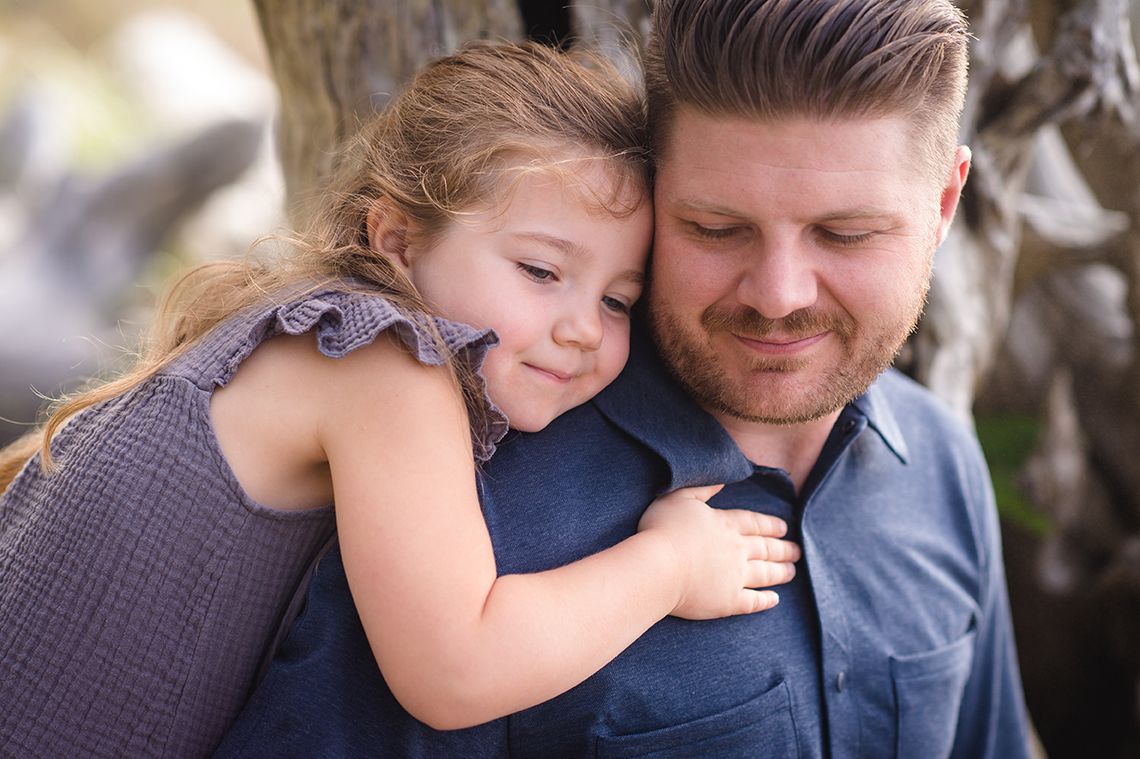 A young child in a gray dress hugs an adult in a blue shirt during a tender outdoor portrait moment.