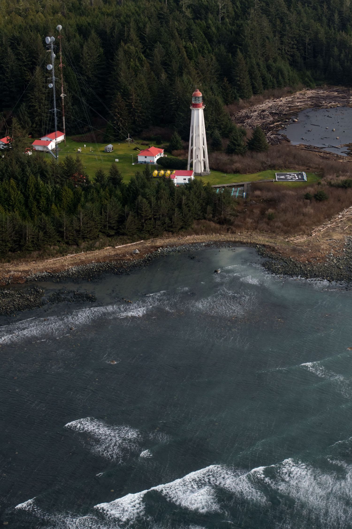 White lighthouse with red roofs stands on a rocky coastline surrounded by evergreen forest and choppy waters.