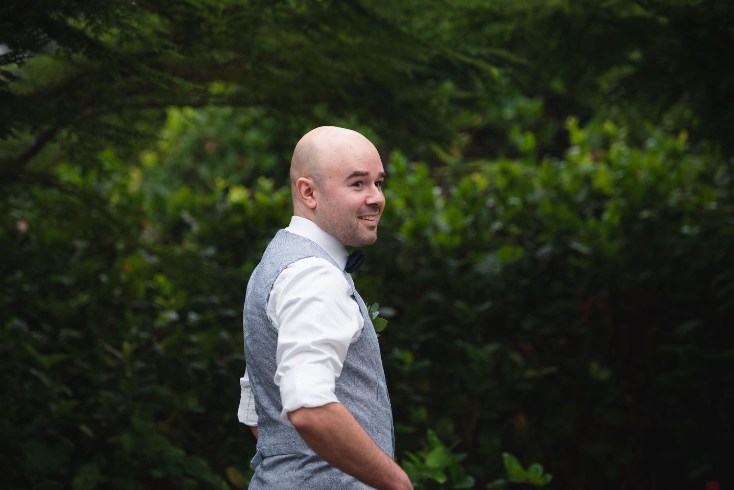 A person in a white dress shirt and gray vest stands outdoors against a green garden background.