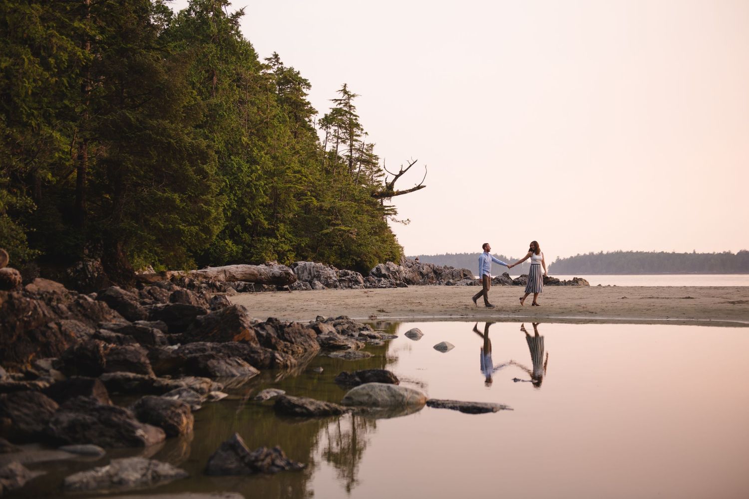 A couple walks hand in hand along a rocky shoreline with calm water and distant islands in view.