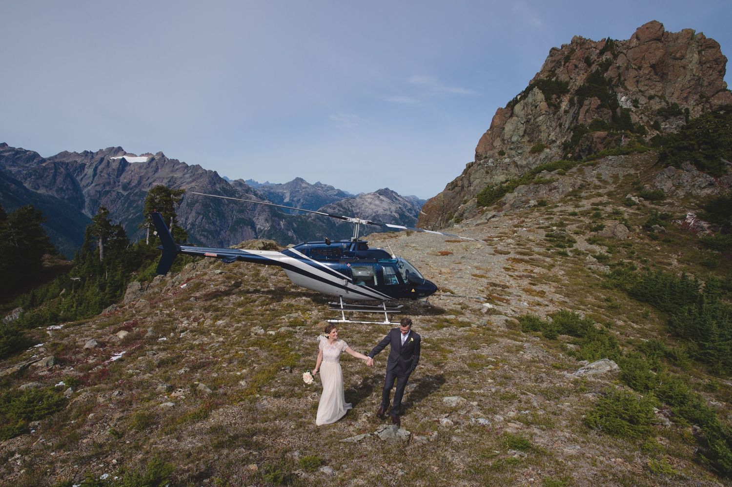 Helicopter landing on a rocky mountain peak with dramatic mountain ranges in the background.