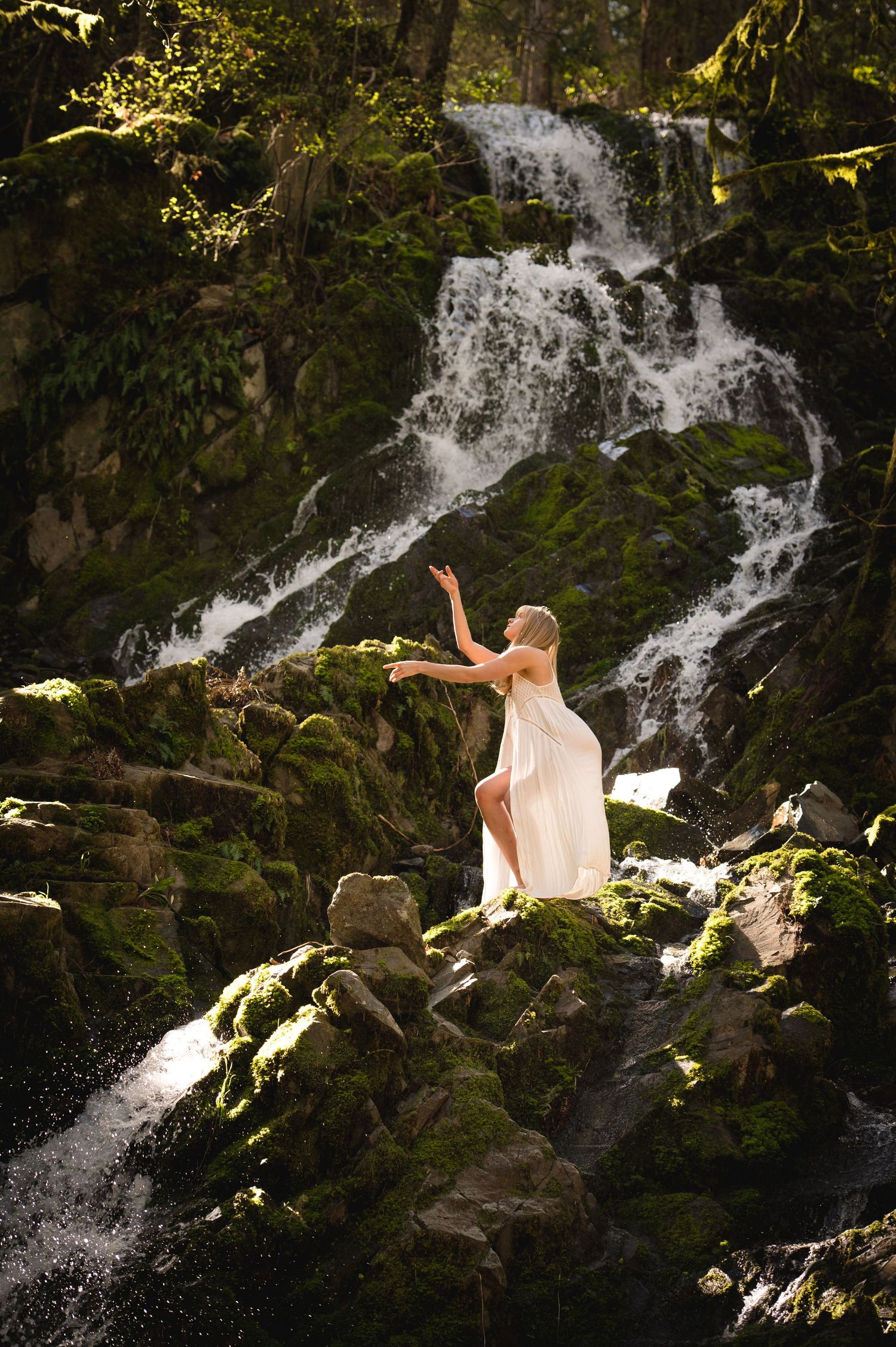 Figure in flowing white dress poses dramatically against a cascading waterfall.