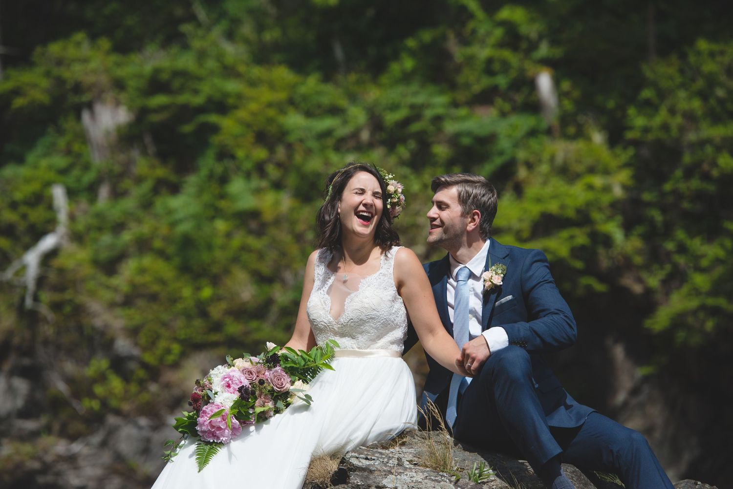 A couple shares a joyful moment while sitting on coastal rocks surrounded by forest.