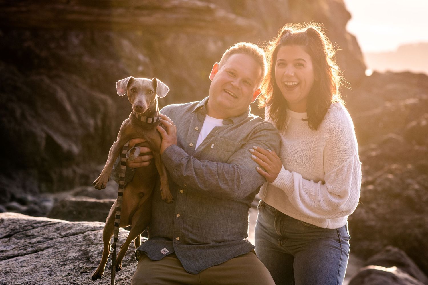 A joyful couple poses on coastal rocks with their small brown dog at sunset.