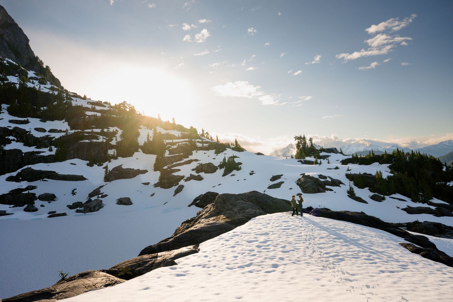 Couple standing on a snowy mountain summit bathed in golden sunset light with panoramic alpine views.