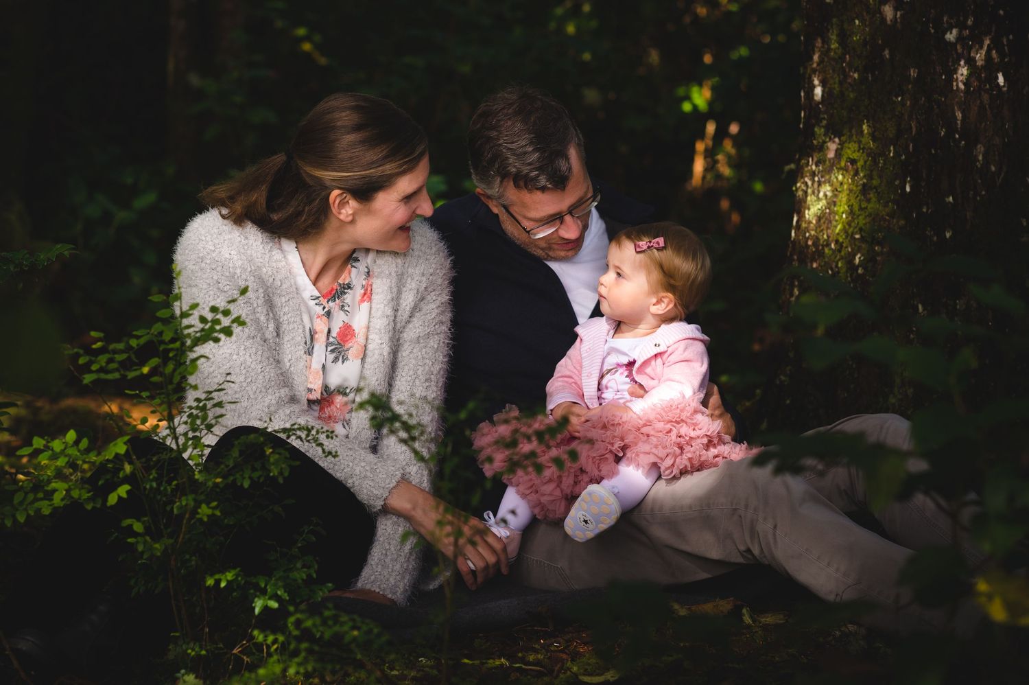 A romantic couple sits together in a sunlit forest surrounded by lush greenery and dappled sunlight.