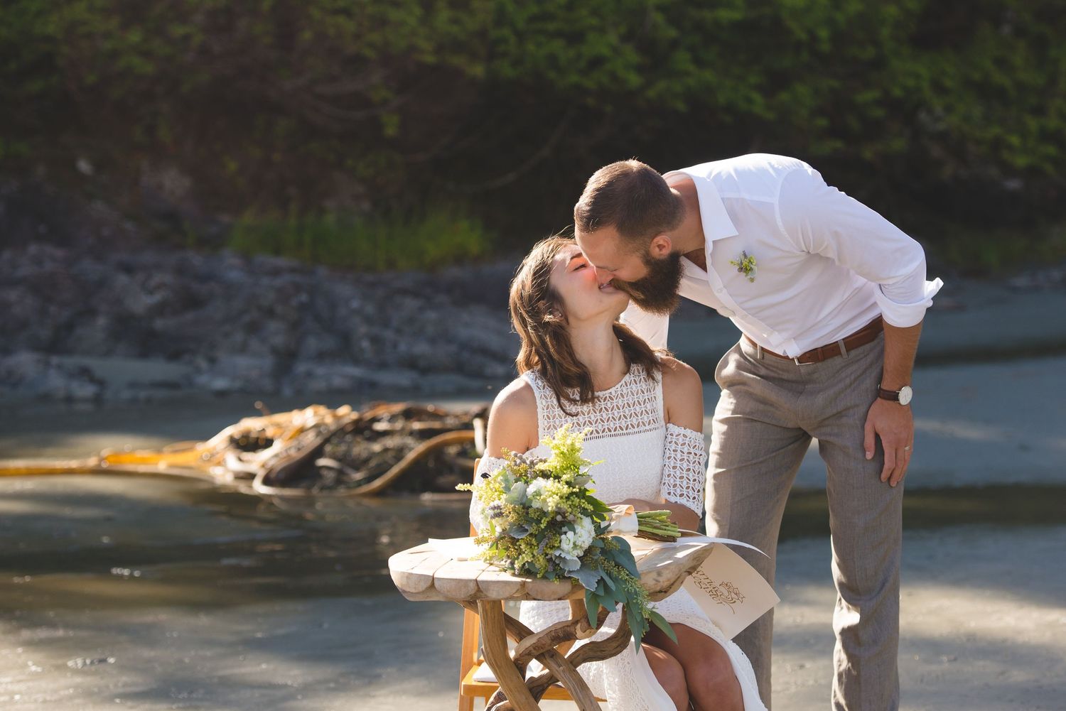 A couple shares a romantic moment by a waterfront with a bouquet of flowers on a rustic table.